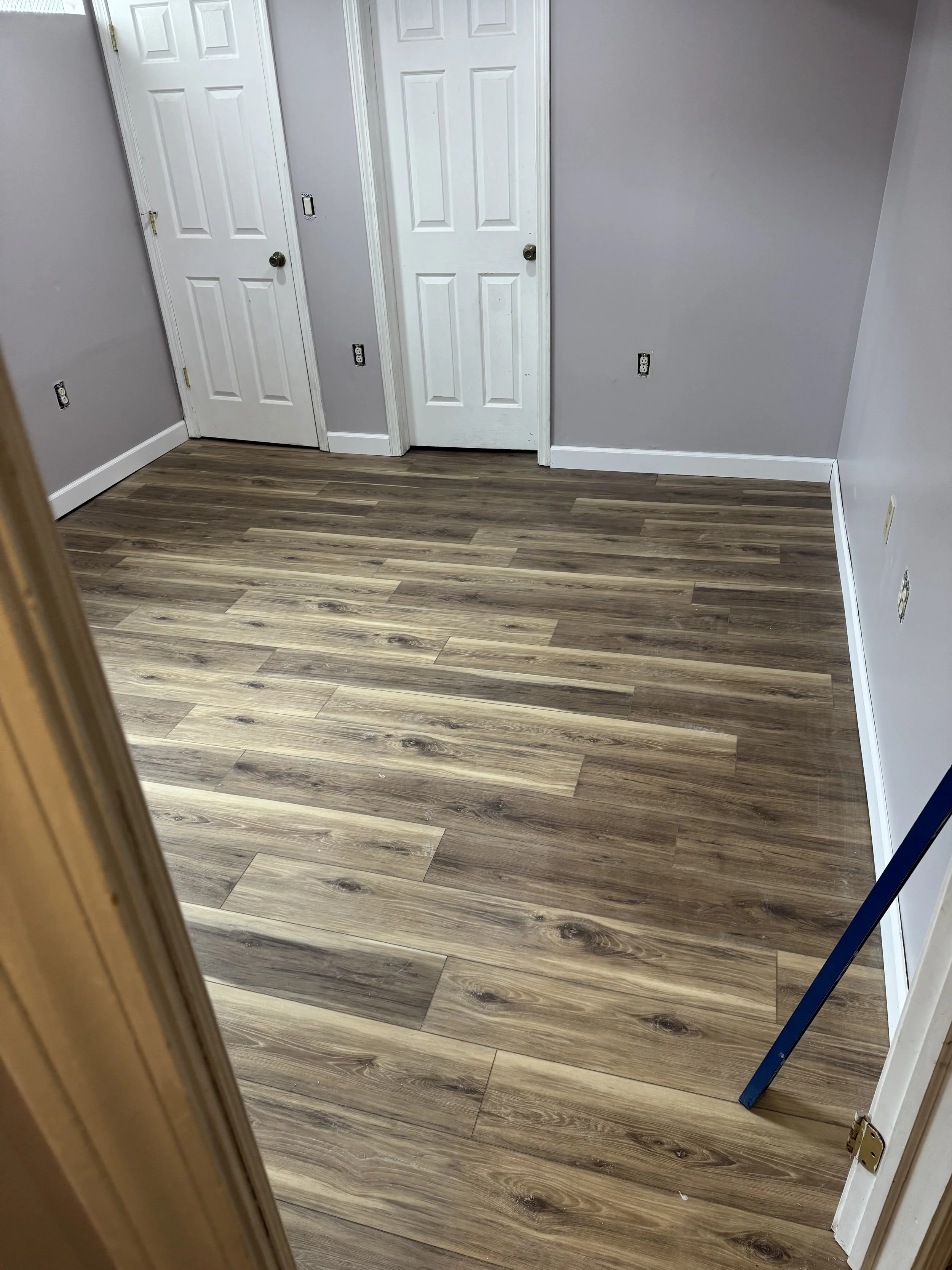 Empty room with gray walls, white trim, and wood-look laminate flooring. There are two white doors, an electrical outlet, and a blue level leaning against the door frame.