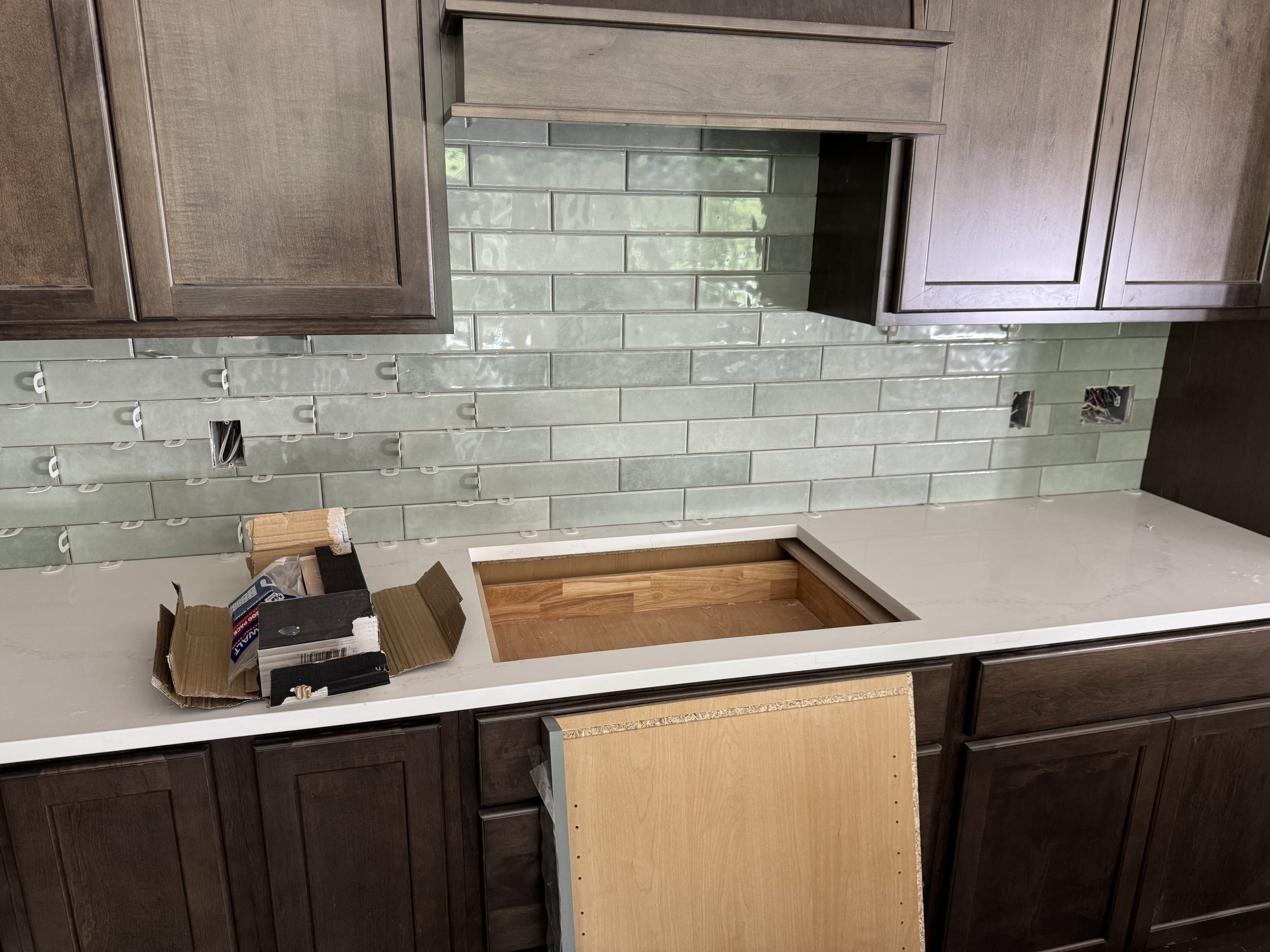 Kitchen with dark wooden cabinets, green subway tile backsplash, and a white countertop. An empty space in the counter is visible, possibly for a sink or appliance. A cardboard box with miscellaneous items is on the counter, and there are two electri