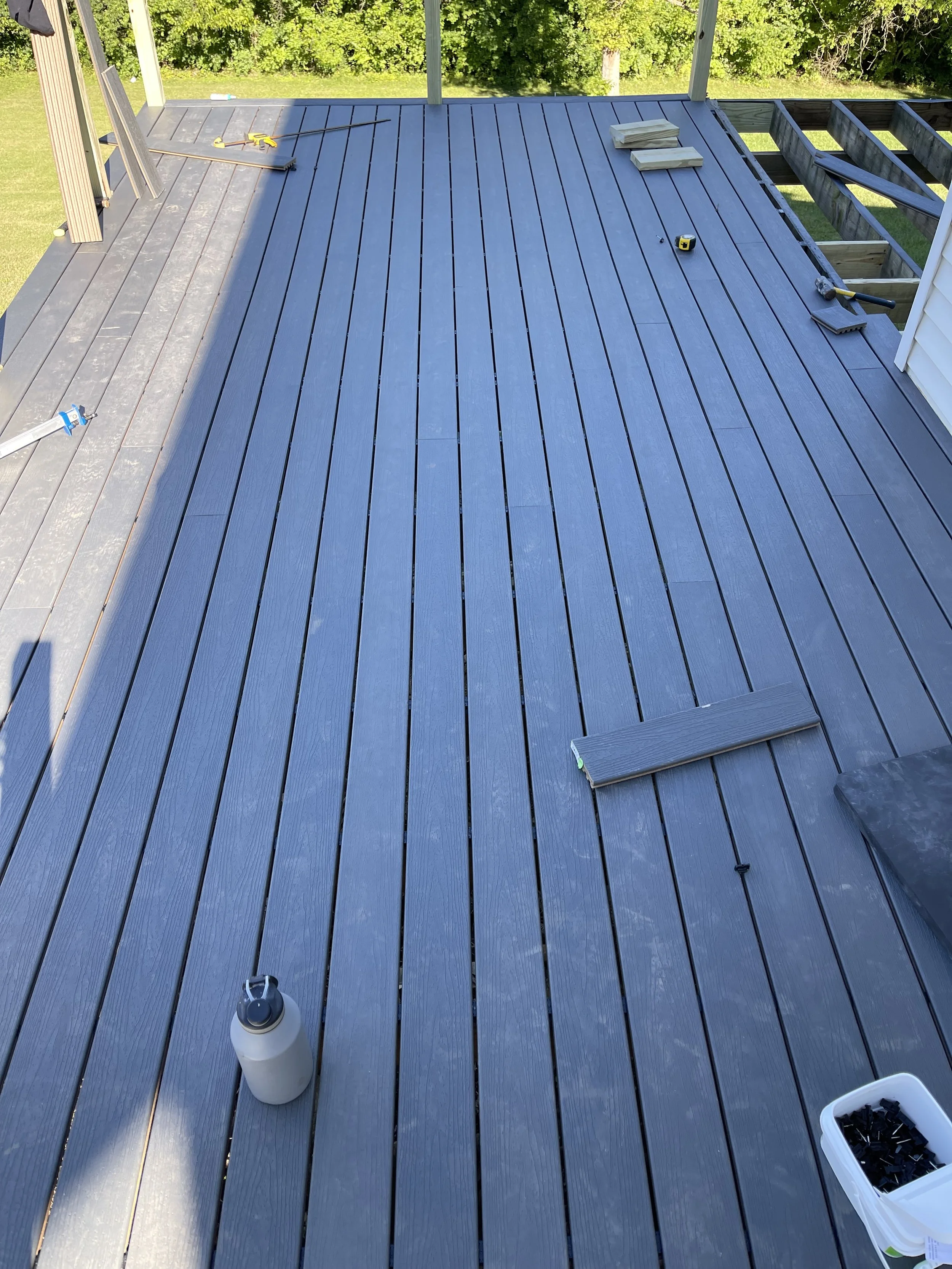 A newly installed dark gray wooden deck outdoors under construction, with tools, wood planks, and a bottle on the surface, surrounded by green trees and a white house siding at the edge.

Installed near oxford Ohio