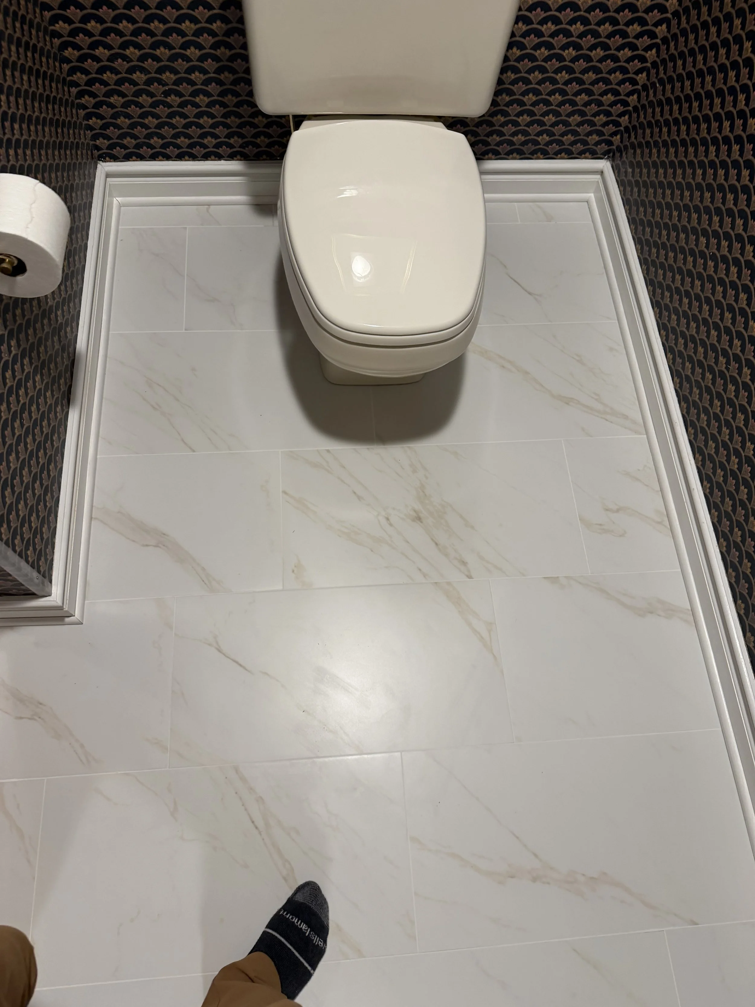 A white ceramic toilet with a closed lid in a bathroom with marble-patterned floor tiles and dark patterned wallpaper. A person's foot is visible in the bottom of the image.