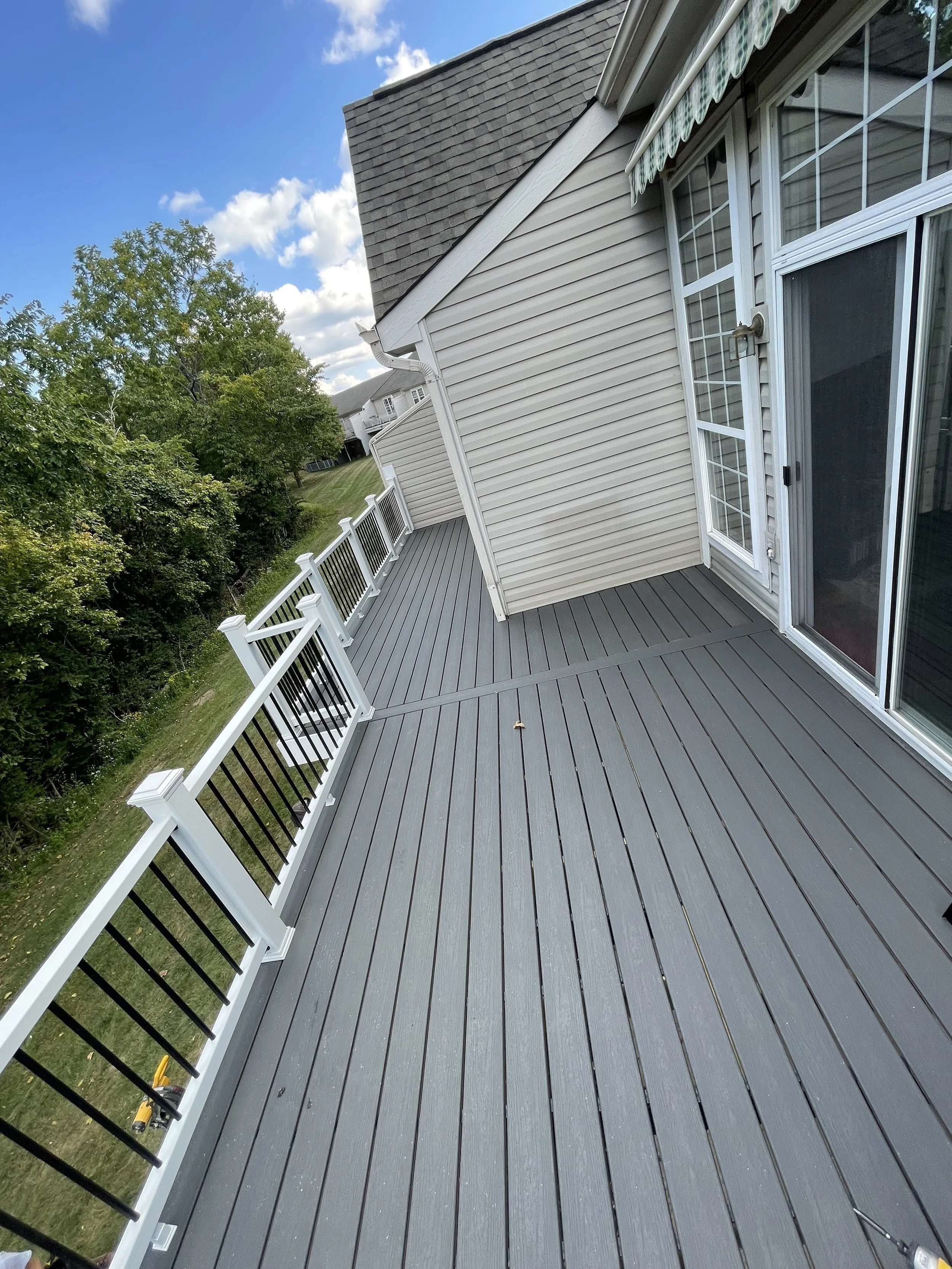 Outside view of a spacious backyard deck with gray flooring, white railing with black balusters, adjacent to a house with white vinyl siding and large windows. Green trees and a partly cloudy blue sky are visible in the background.