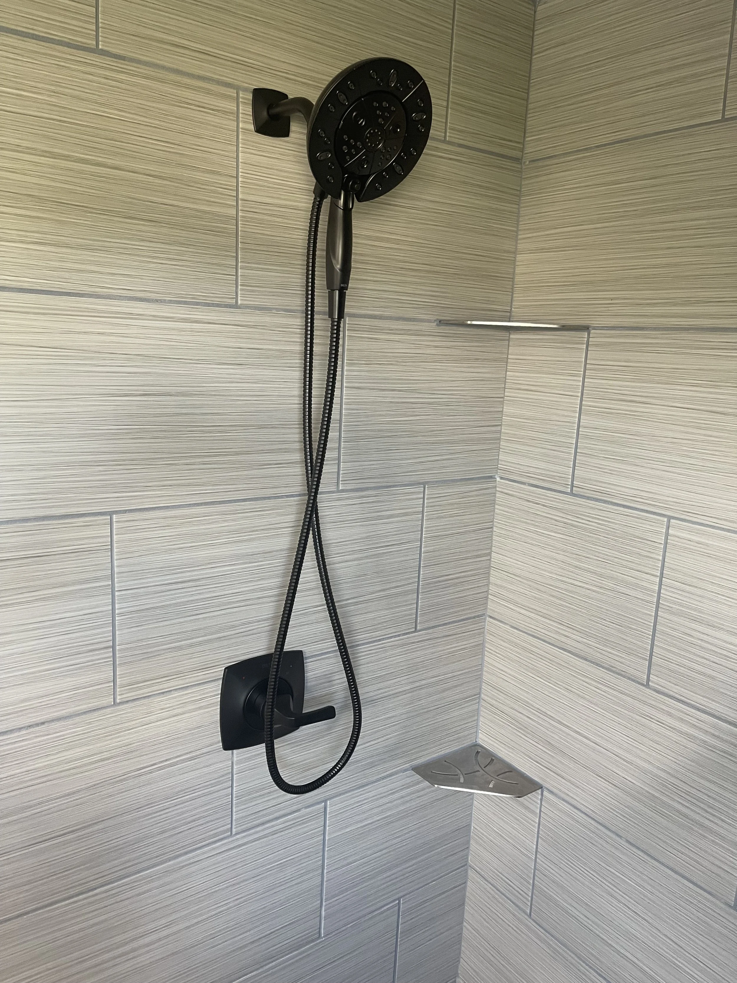 A black shower head with a handheld hose mounted on a tiled bathroom wall.