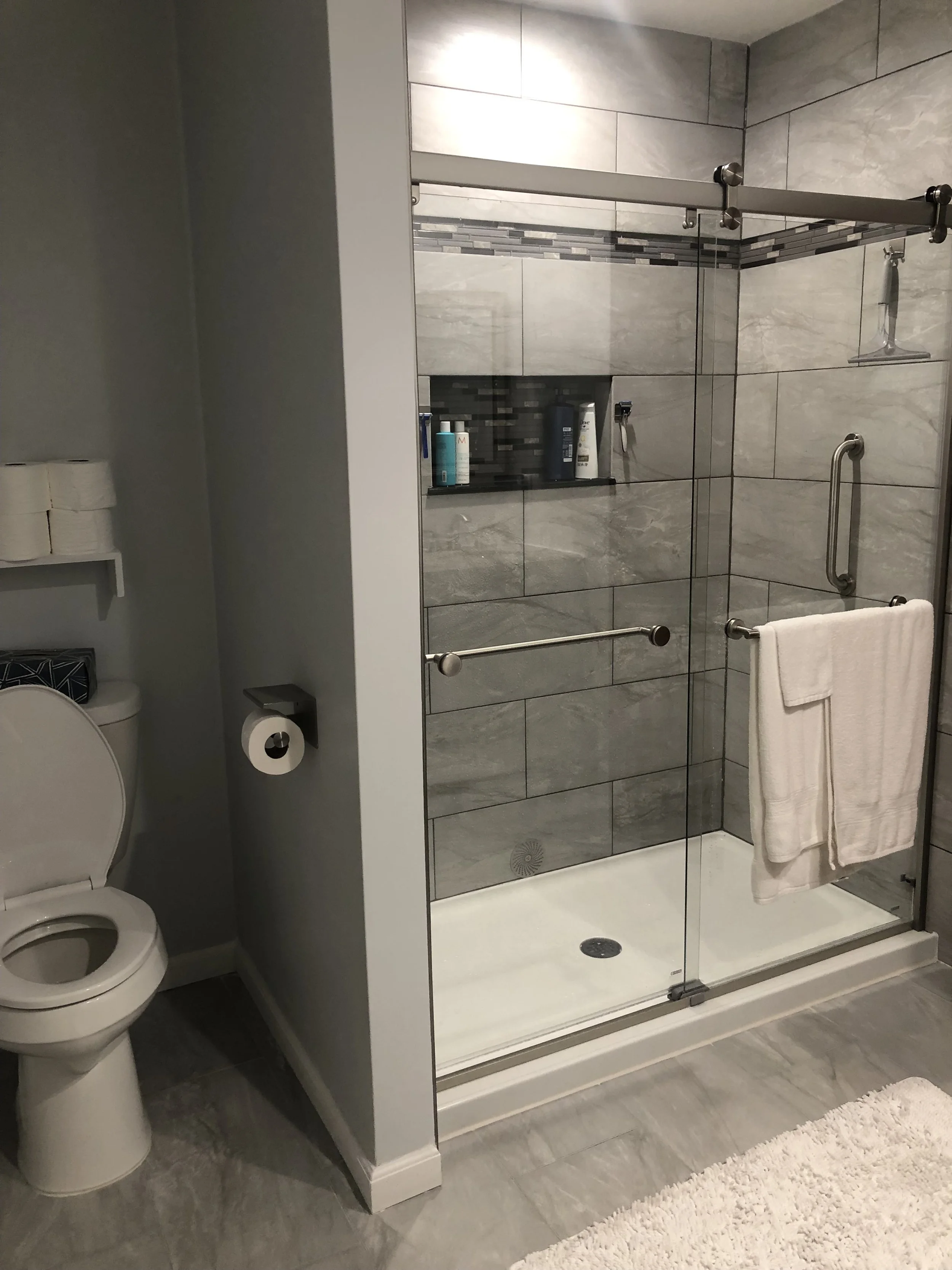 A bathroom with a glass-enclosed shower featuring gray tile walls and a built-in shelf with toiletries. To the left, there is a toilet with a roll of toilet paper on a wall-mounted holder. The floor and surrounding area have gray tiles, and a white b
