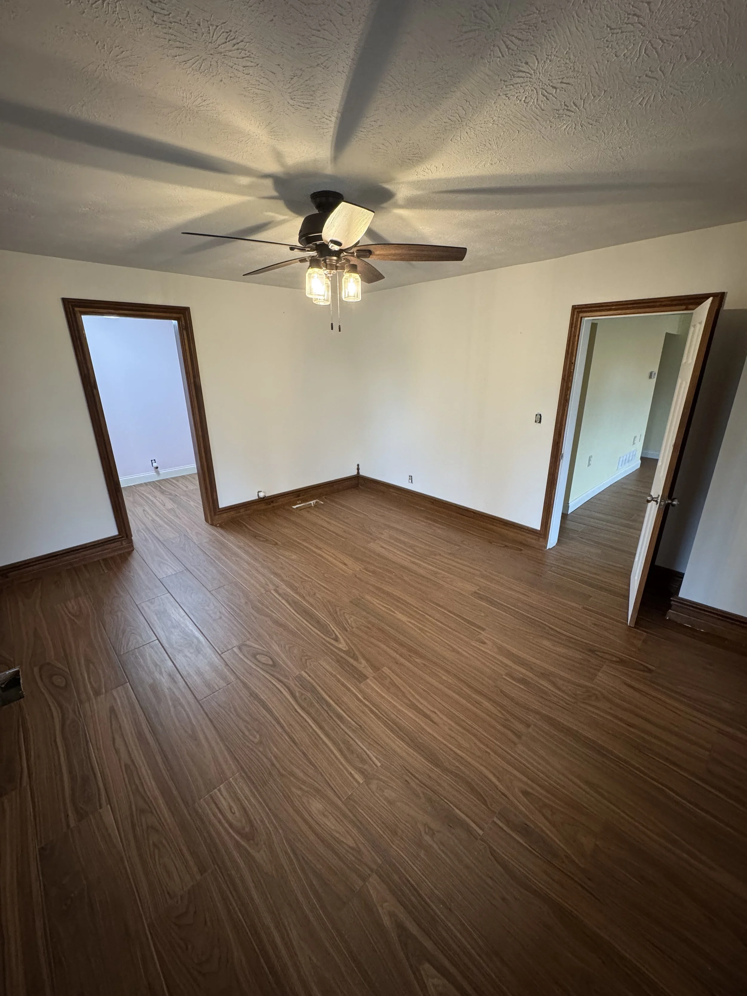 Empty room with light-colored walls, wood flooring, a ceiling fan with lights, and two doorways leading to other rooms.