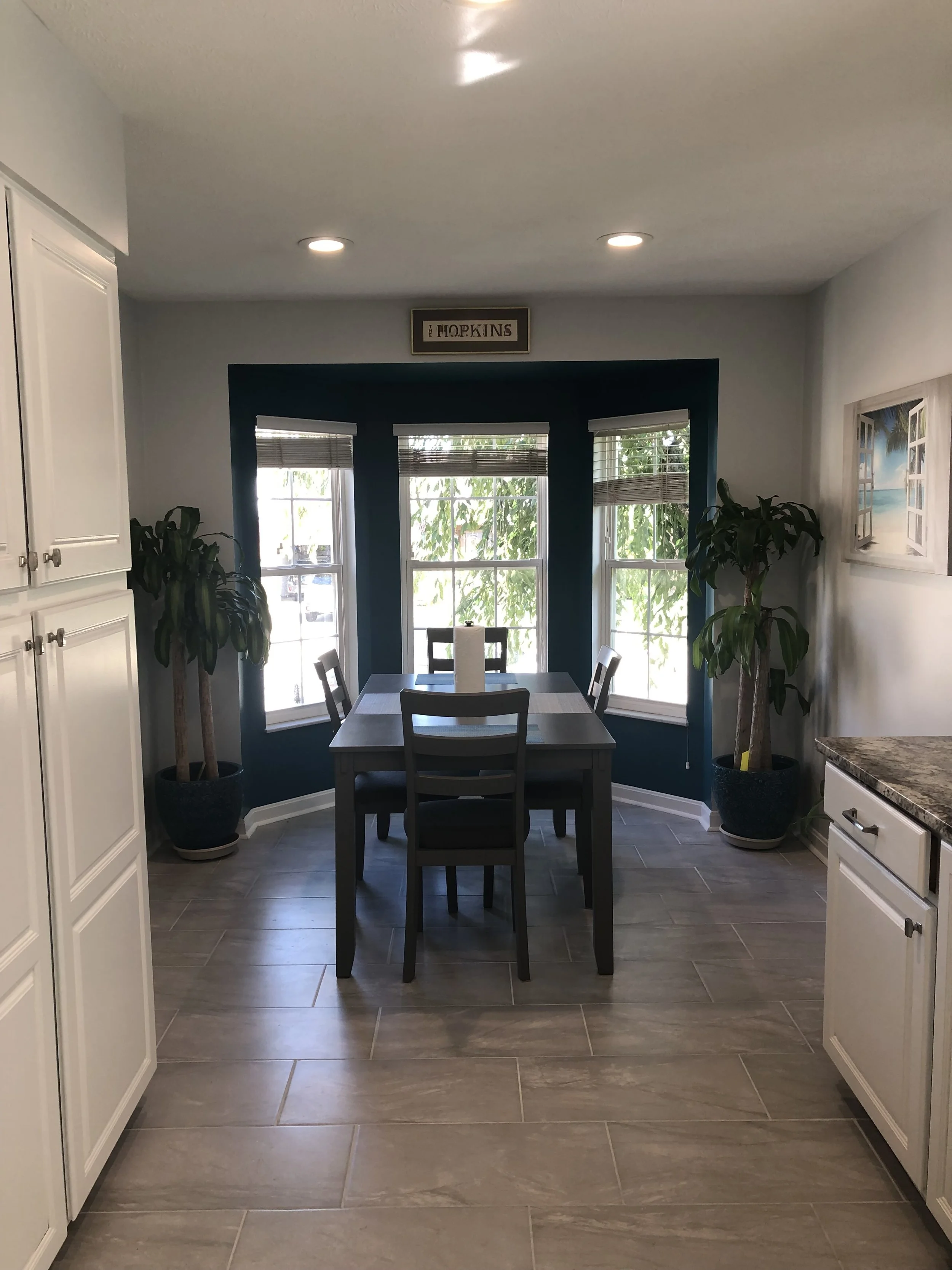 Dining area with a dark wooden table and four matching chairs, situated near a bay window with three sections, all with blinds. Two large potted plants are flanking the window, and a picture with an open window view hangs on the wall to the right. Th