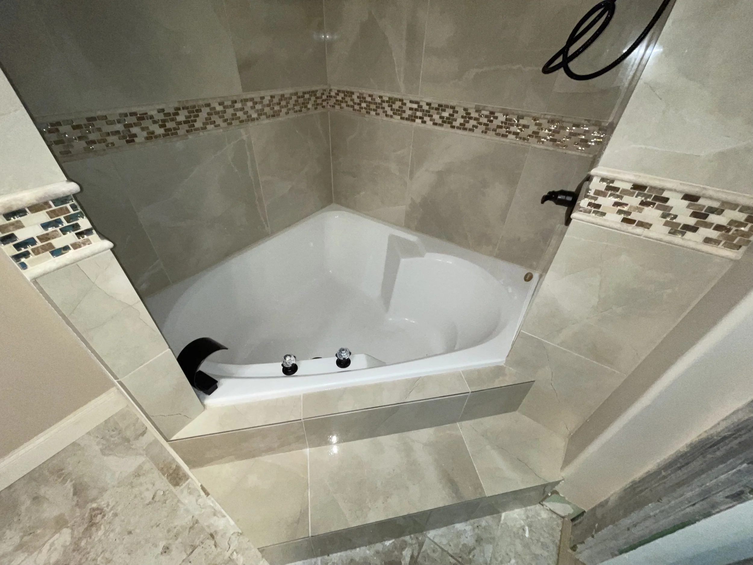 Bathroom corner with a tiled wall, corner bathtub with black faucet handle, and decorative tile border.
