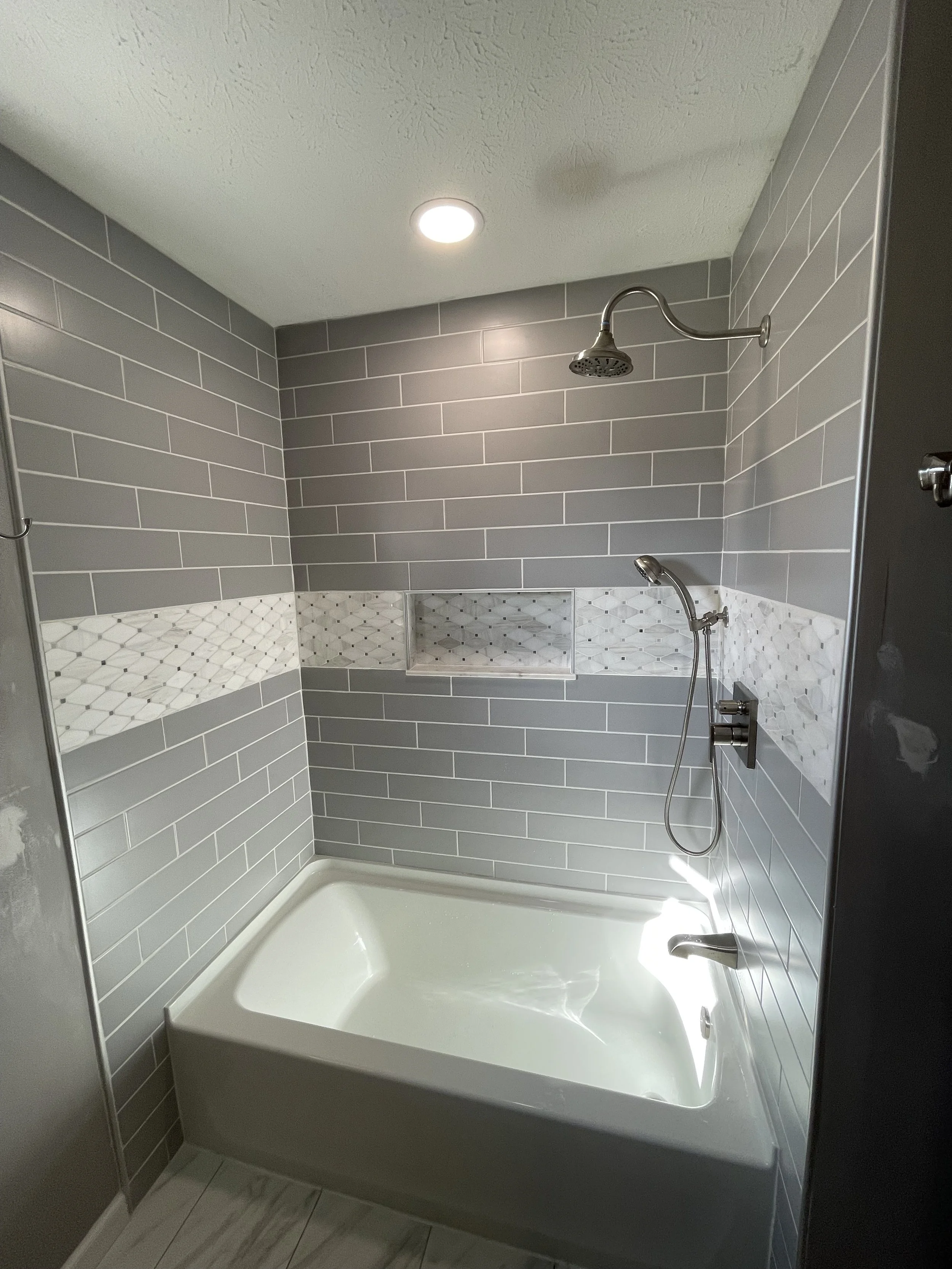 Modern bathroom shower with gray subway tiles, decorative tile strip, bathtub, overhead shower head, and handheld shower.