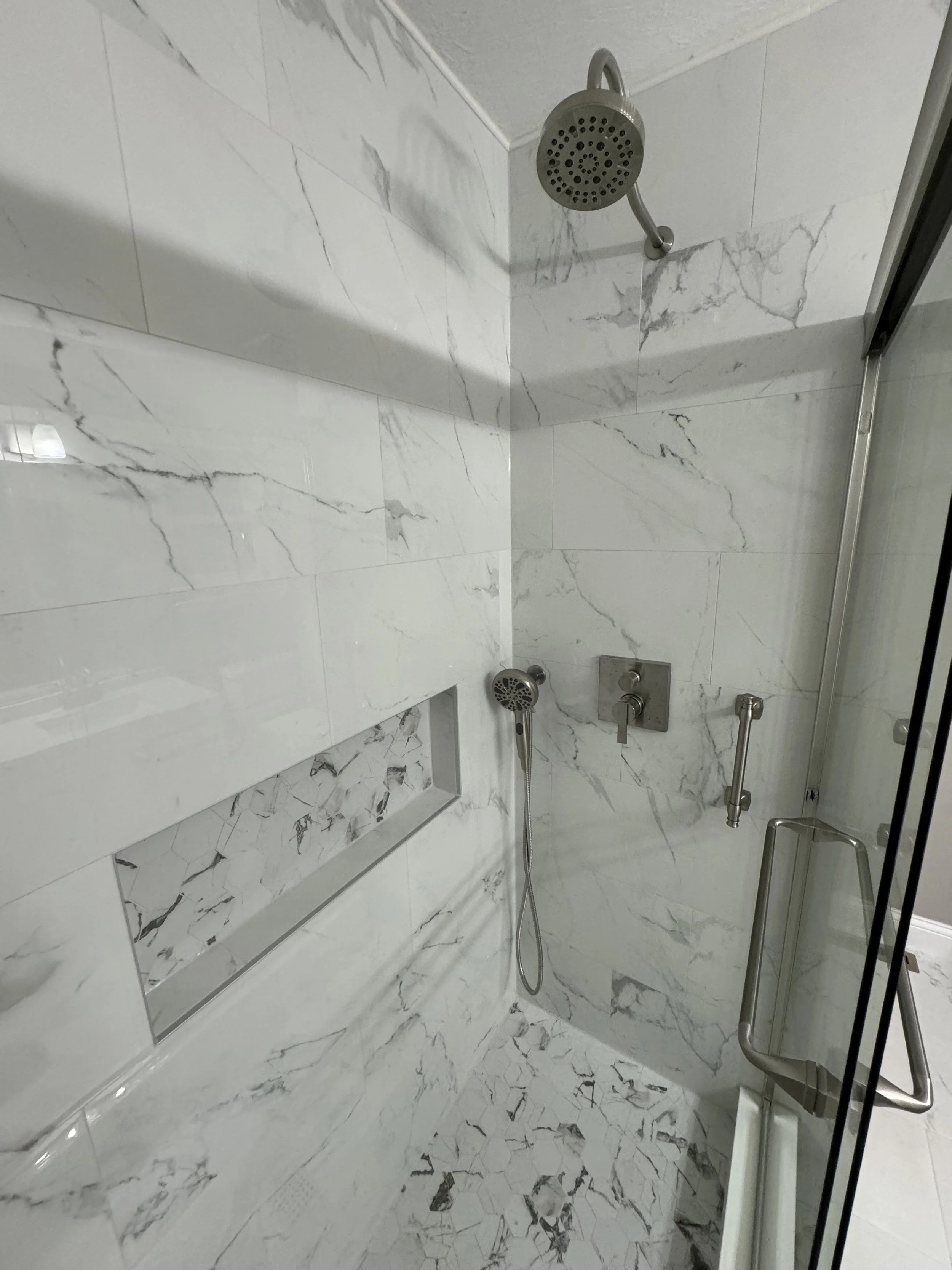 White marble tiled walk-in shower with a handheld showerhead, large overhead shower, built-in niche for toiletries, and a glass door.