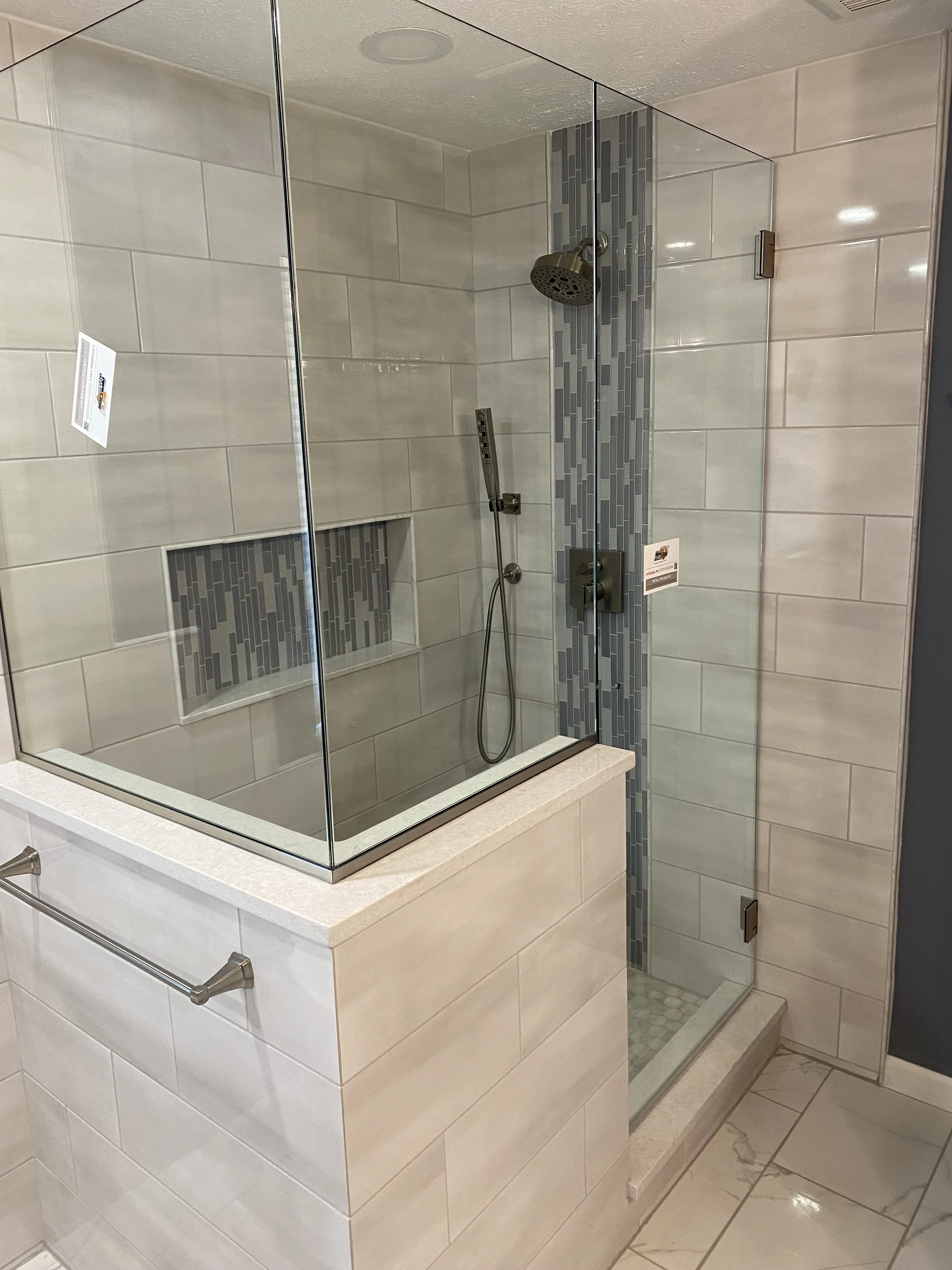A modern walk-in shower with beige tiled walls, a glass enclosure, a built-in tile niche, and a vertical strip of gray mosaic tiles. It features a rainfall showerhead and a handheld shower attachment.