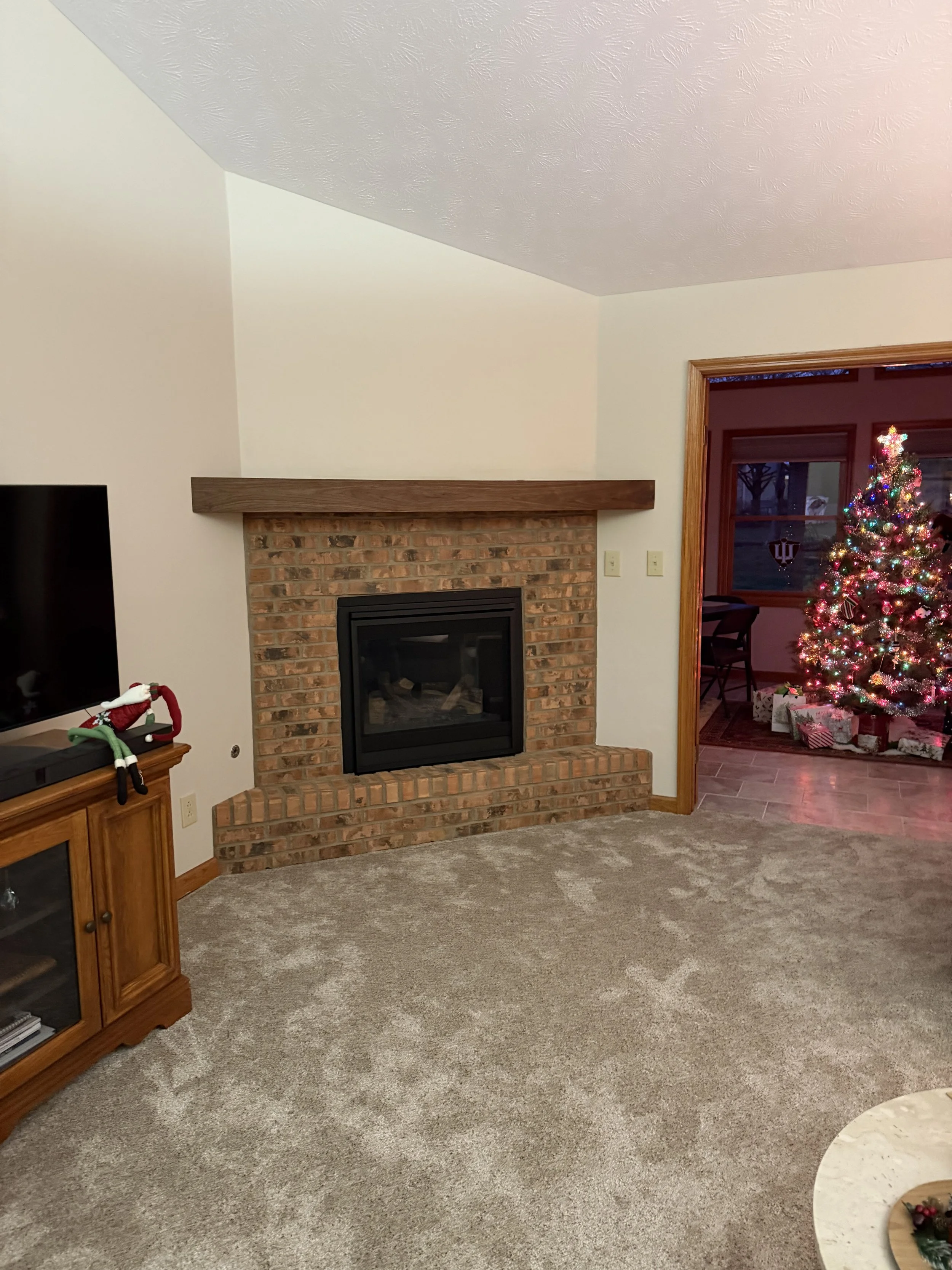 Living room with a brick fireplace, a TV with a Santa hat on it, and a Christmas tree with presents underneath in the background.