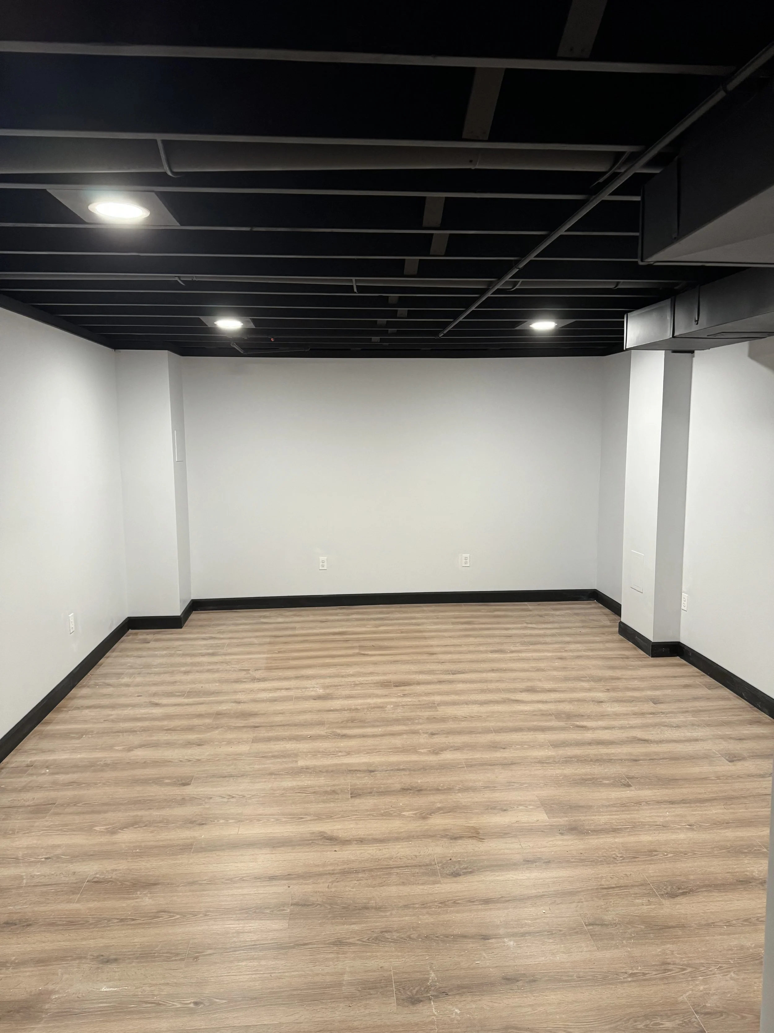 Empty room with light wood flooring, white walls, black baseboards, and a black ceiling with track lighting.
