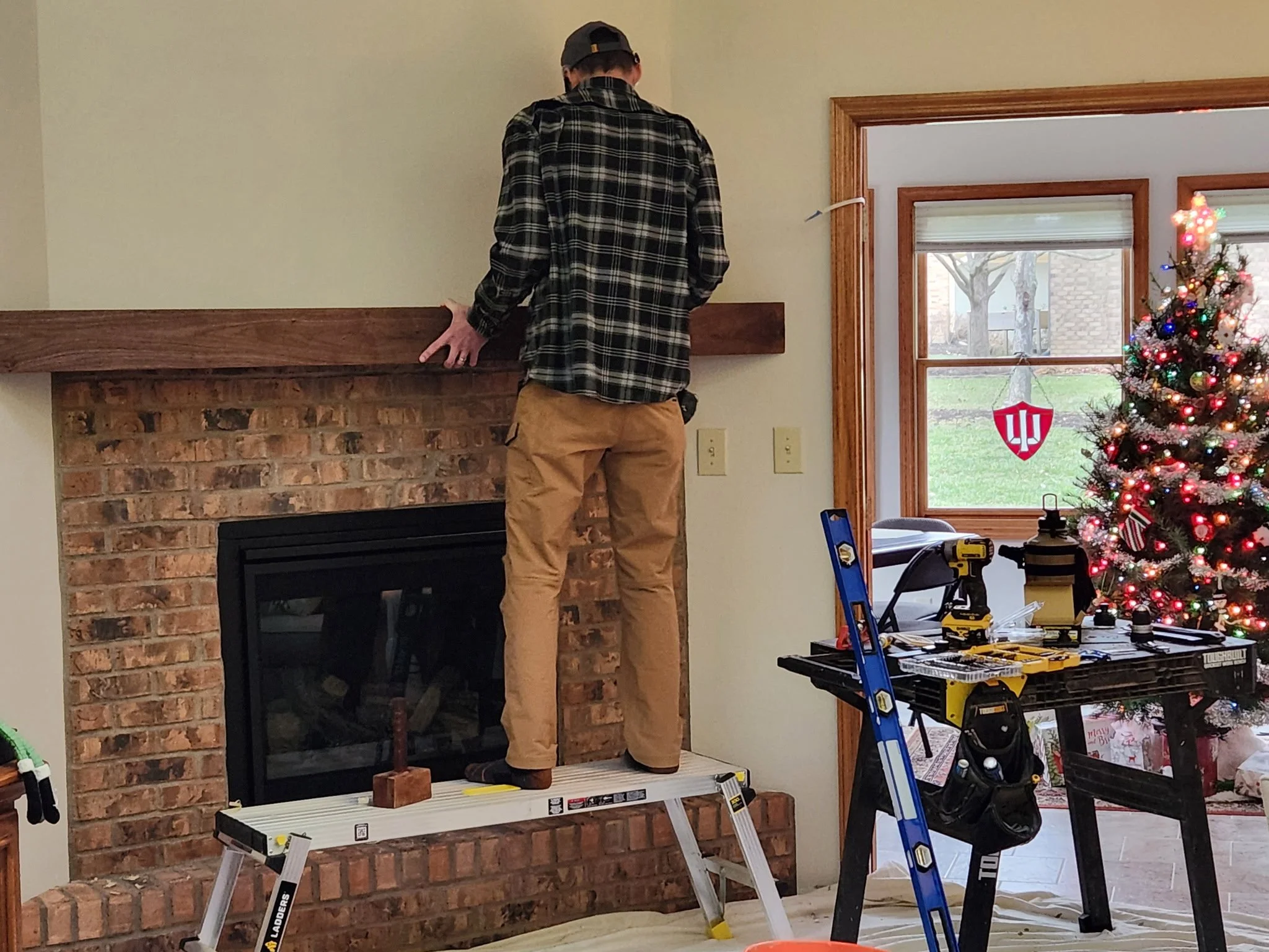 A person on a step ladder working on a brick fireplace mantel in a living room, with Christmas decorations and tools around.