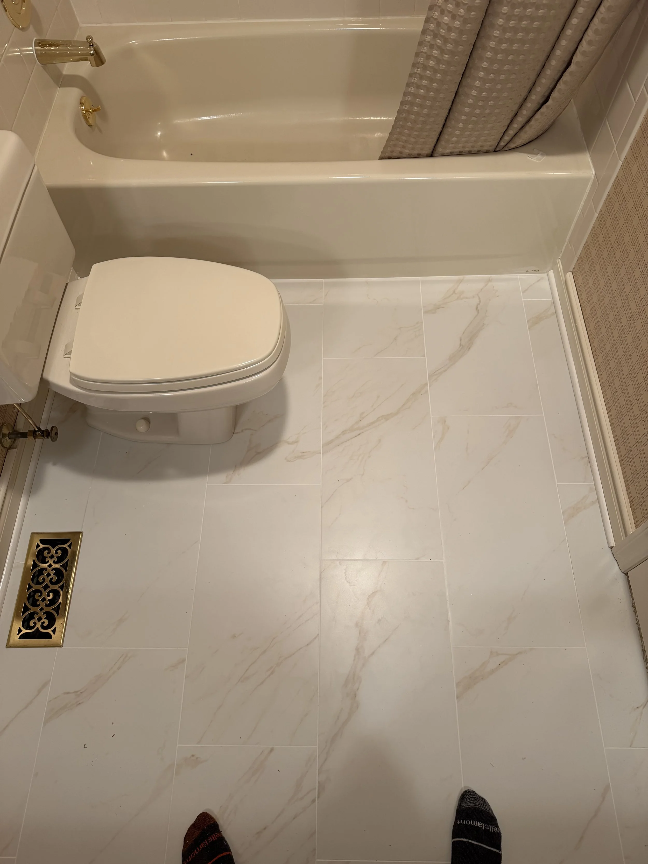 A bathroom with white marble tiles, a toilet, a bathtub with a beige curtain, and a decorative floor vent. The photo is taken from a perspective standing over the person's feet.