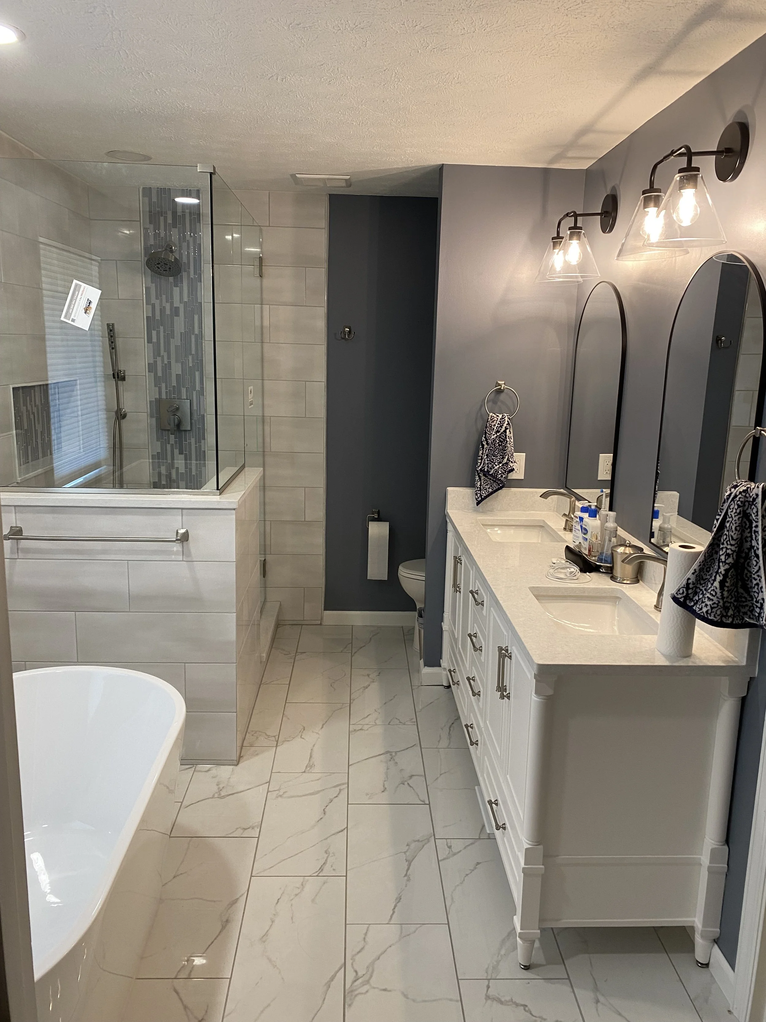 A modern bathroom with a bathtub, a glass-enclosed shower, a toilet, and a white vanity with two sinks and oval mirrors. Four light fixtures are above the vanity. The flooring and shower are tiled with white marble pattern, and the walls are painted 