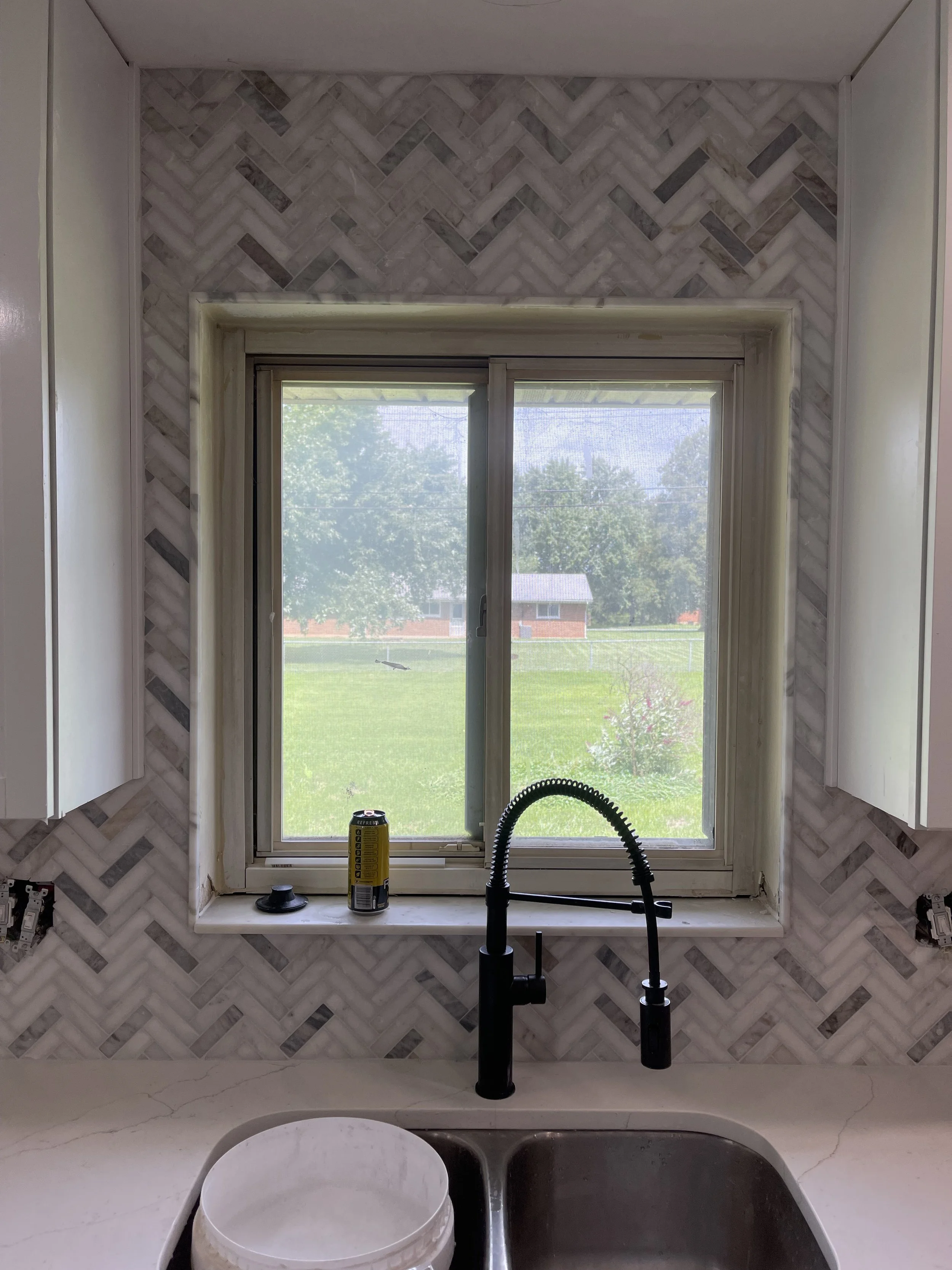 Kitchen sink with black faucet, window with view of a yard, and a yellow can on the windowsill, with a white bucket in the sink.