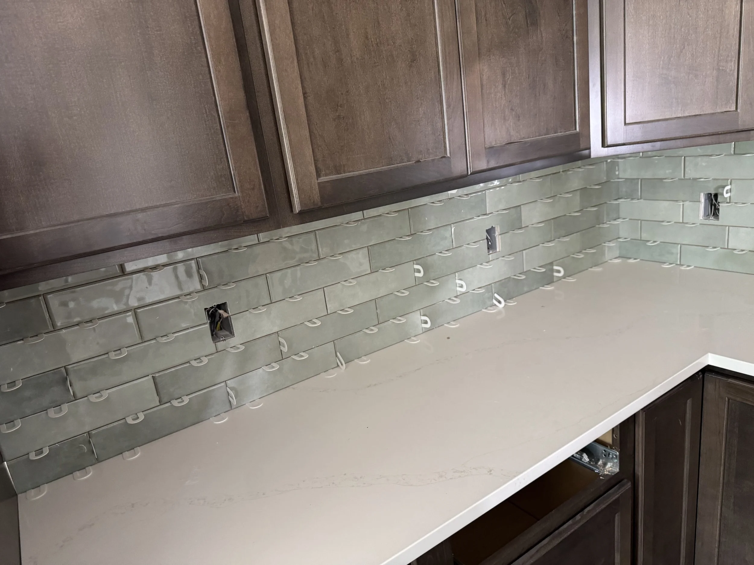 Kitchen backsplash under construction with green subway tiles and exposed electrical outlets, dark wooden cabinets above, and light-colored countertop.
