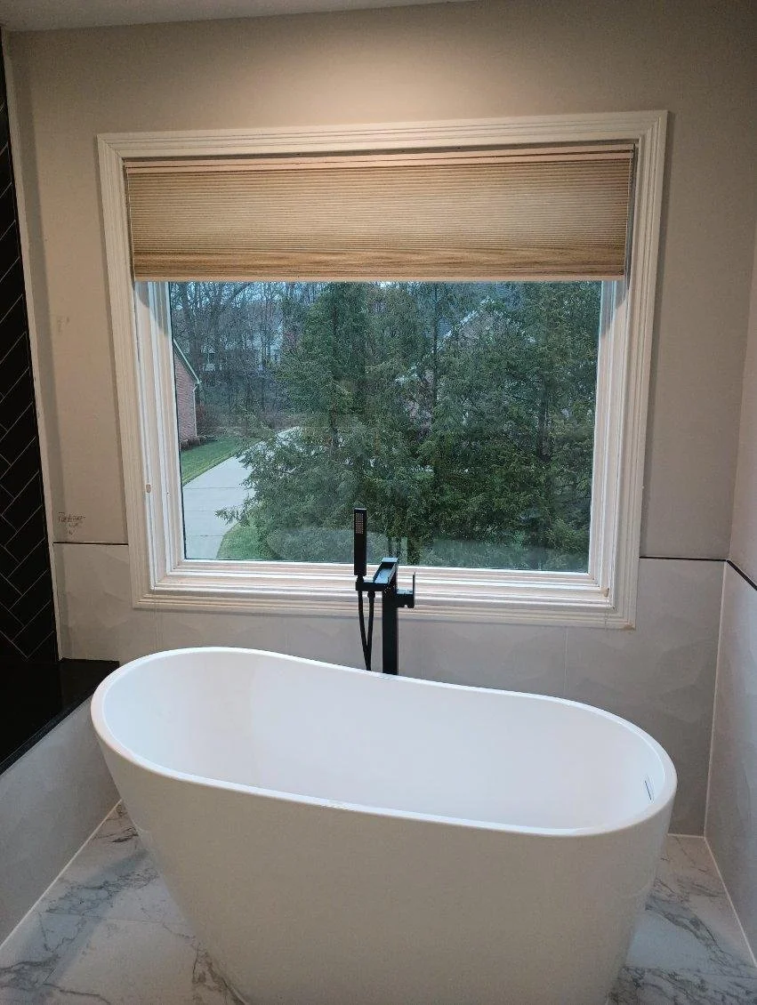 Modern white bathtub with black faucet, large window with beige shade, view of trees and neighborhood outside.