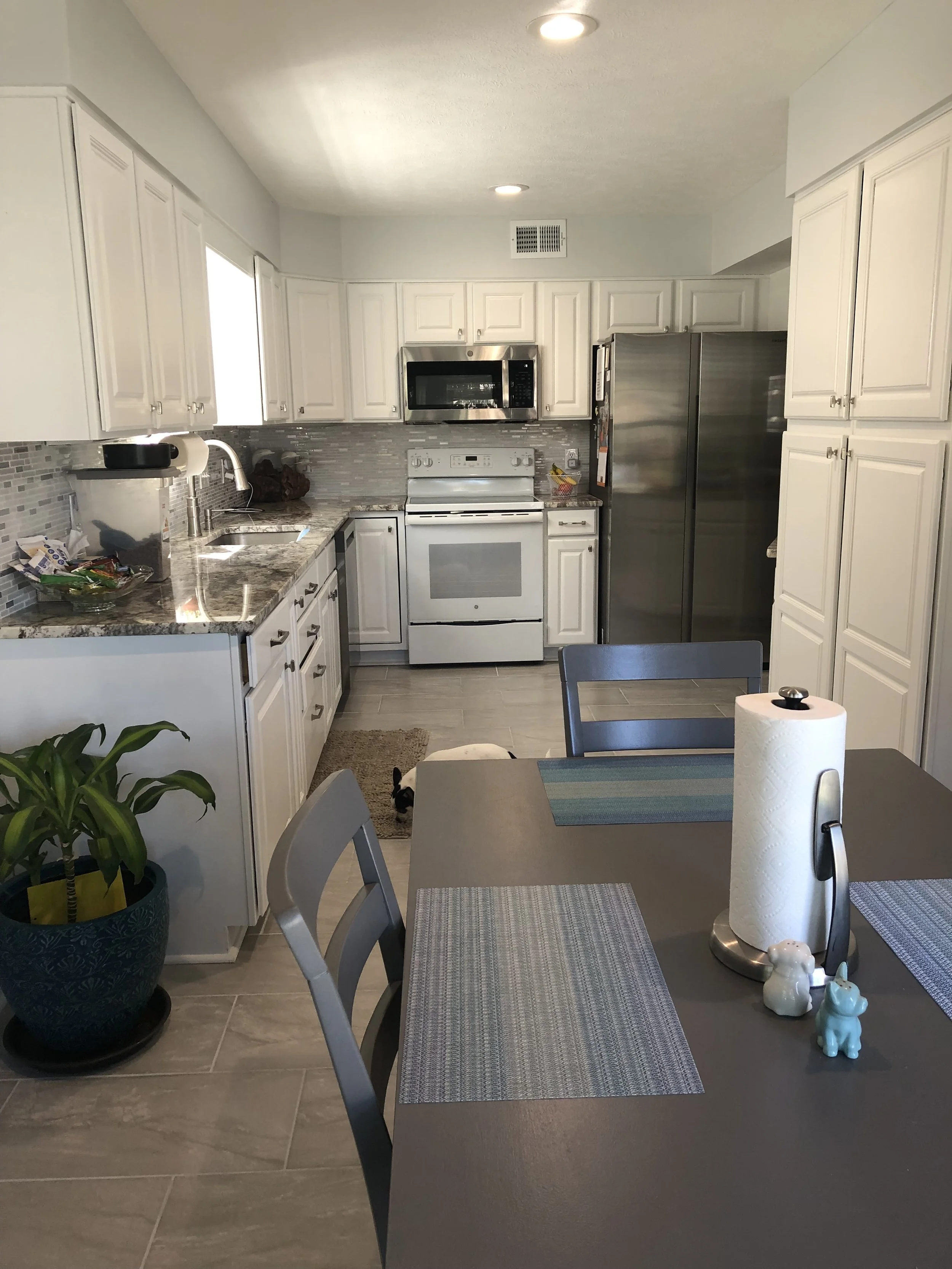 A kitchen with white cabinets, stainless steel appliances, a granite countertop, and a dining table with placemats, a paper towel roll, and decorative items. A small dog is visible under the table.