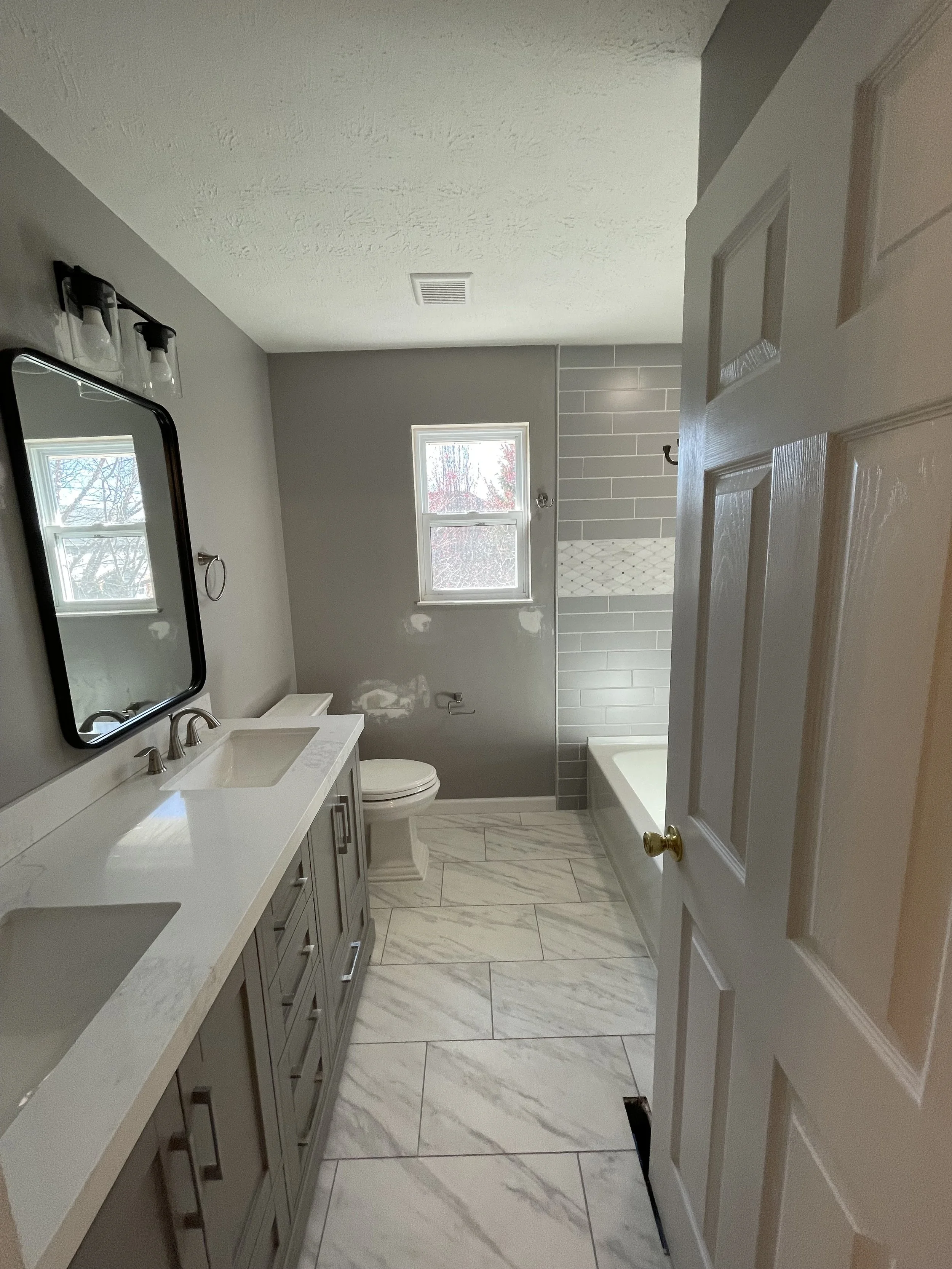 A bathroom with a gray vanity, white countertop, and two sinks. There is a mirror above the vanity with three light fixtures. The bathroom has a toilet, a window, and a bathtub surrounded by gray and white tile. The floor is tiled with marble-pattern