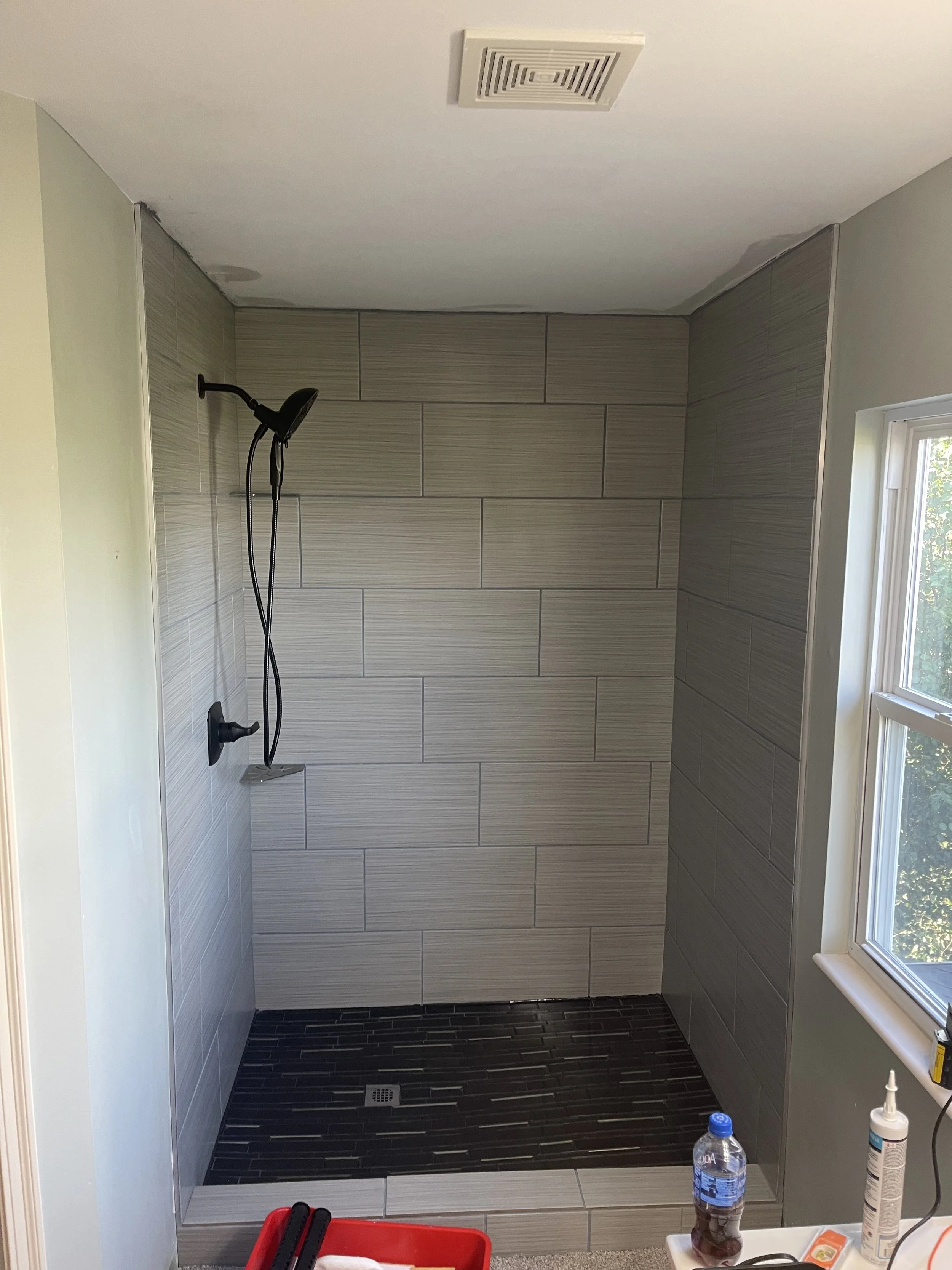 Empty walk-in shower with large gray tiles on the walls, dark rectangular tiles on the floor, a black handheld showerhead, and a small window on the right.


Installed in Cincinnati Ohio 