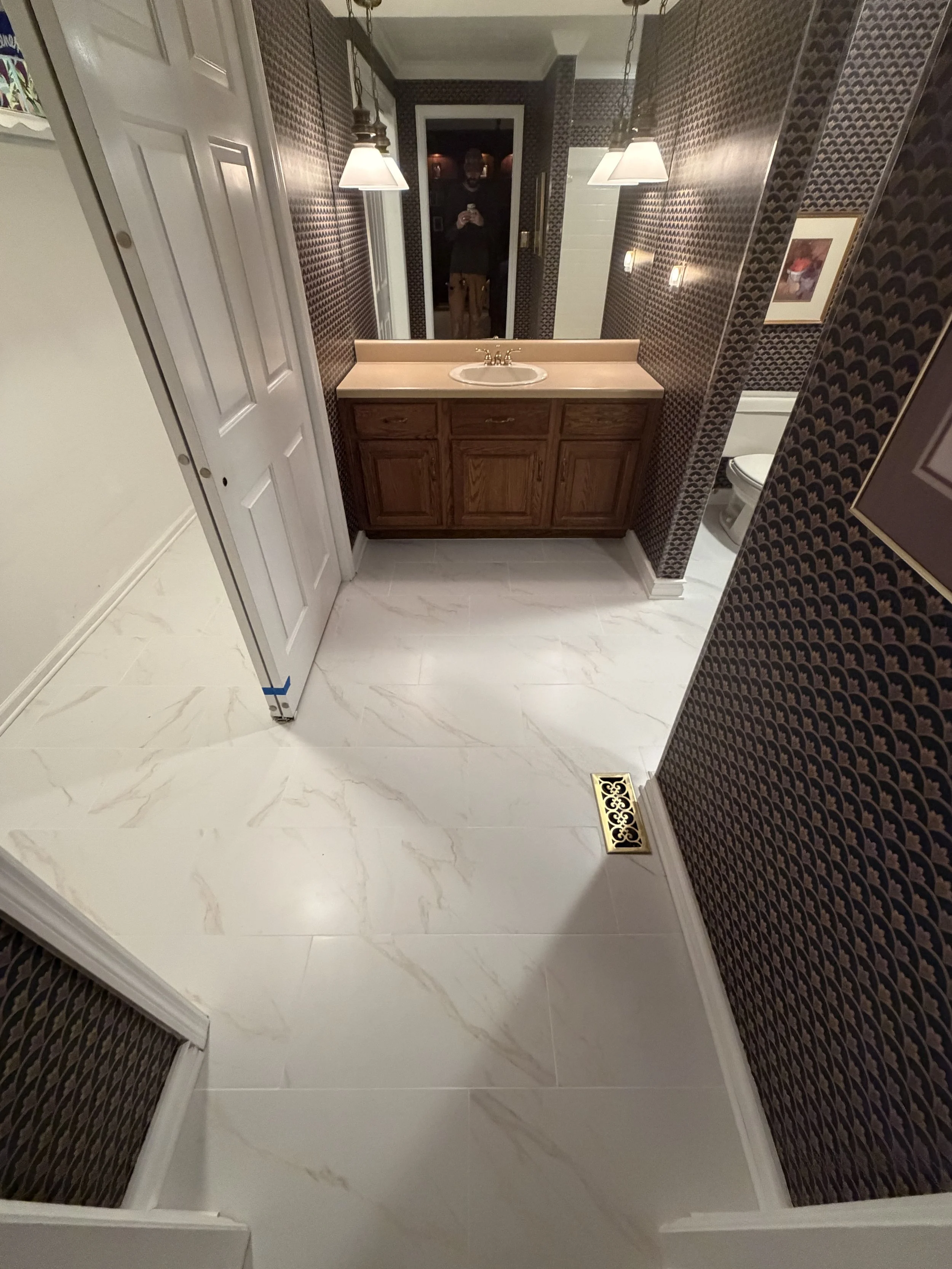 Bathroom with dark-patterned wallpaper, a wooden vanity with a small sink, a mirror above, two wall-mounted lights, a toilet, and a person's reflection in the mirror taking a photo.