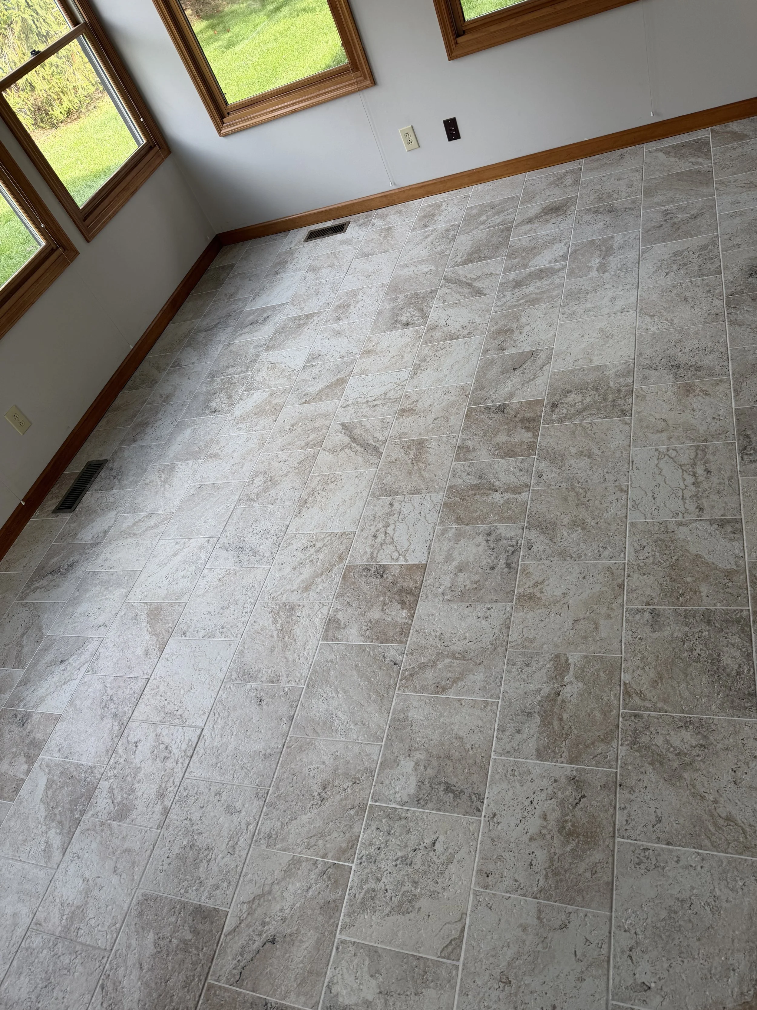 Empty room with tiled beige and gray floor, three windows with wooden frames, and baseboard molding along the wall.