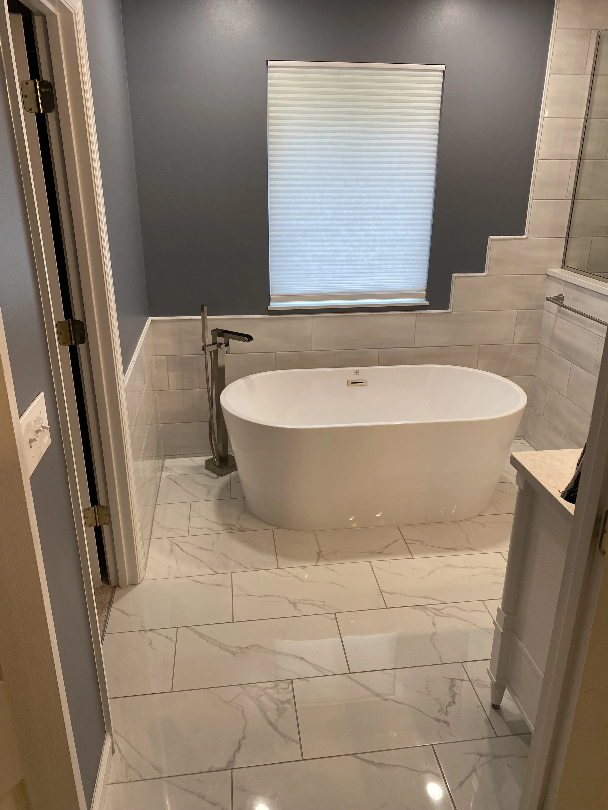 Modern bathroom with a white freestanding bathtub, gray wall, marble tile flooring, window with closed blinds, and a glass shower enclosure. 

Installed Near Mason Ohio