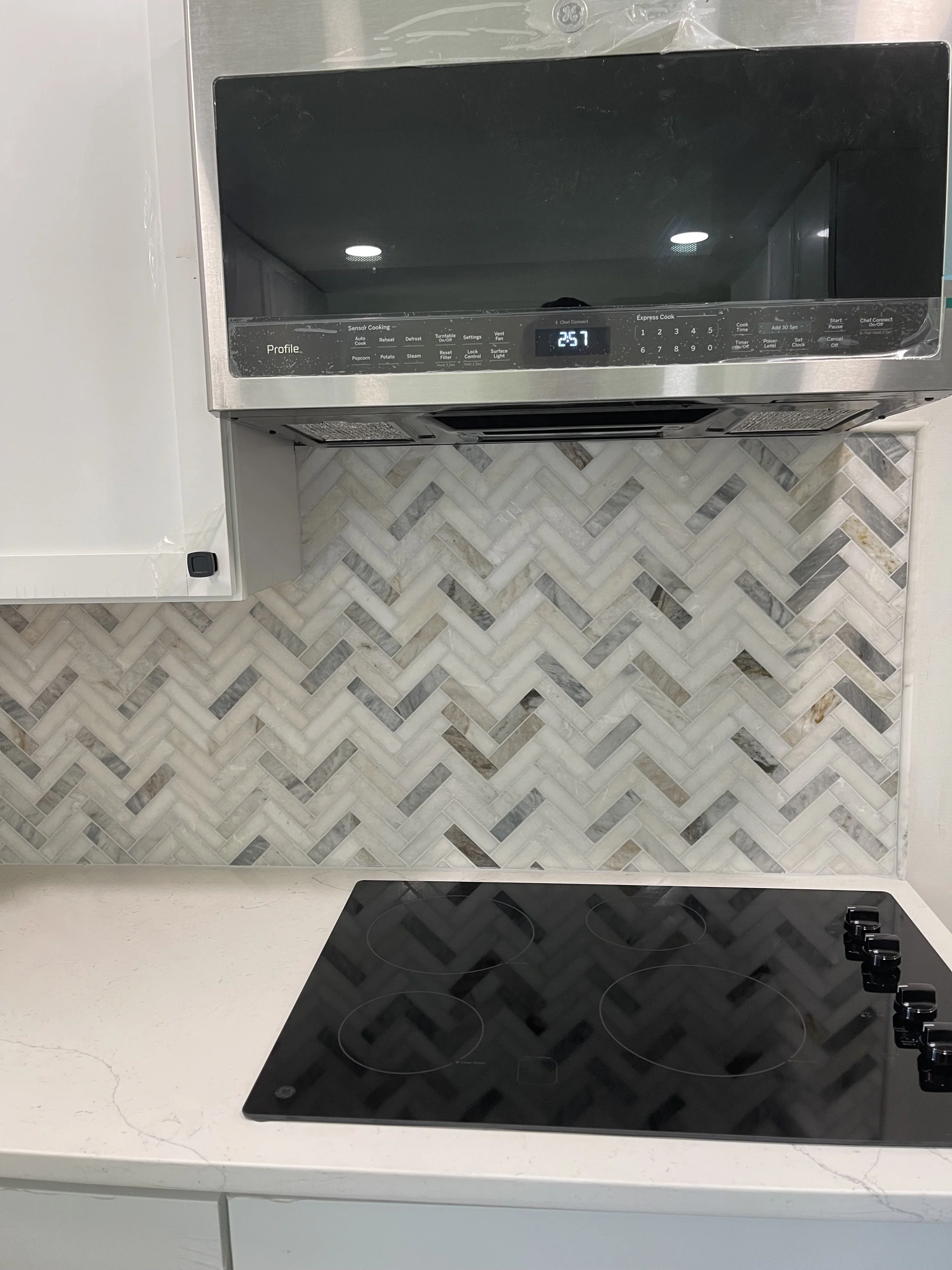 Modern kitchen stove with a black glass top and four burners, situated beneath a stainless steel microwave with a digital display reading 25.7, against a marble tile backsplash.