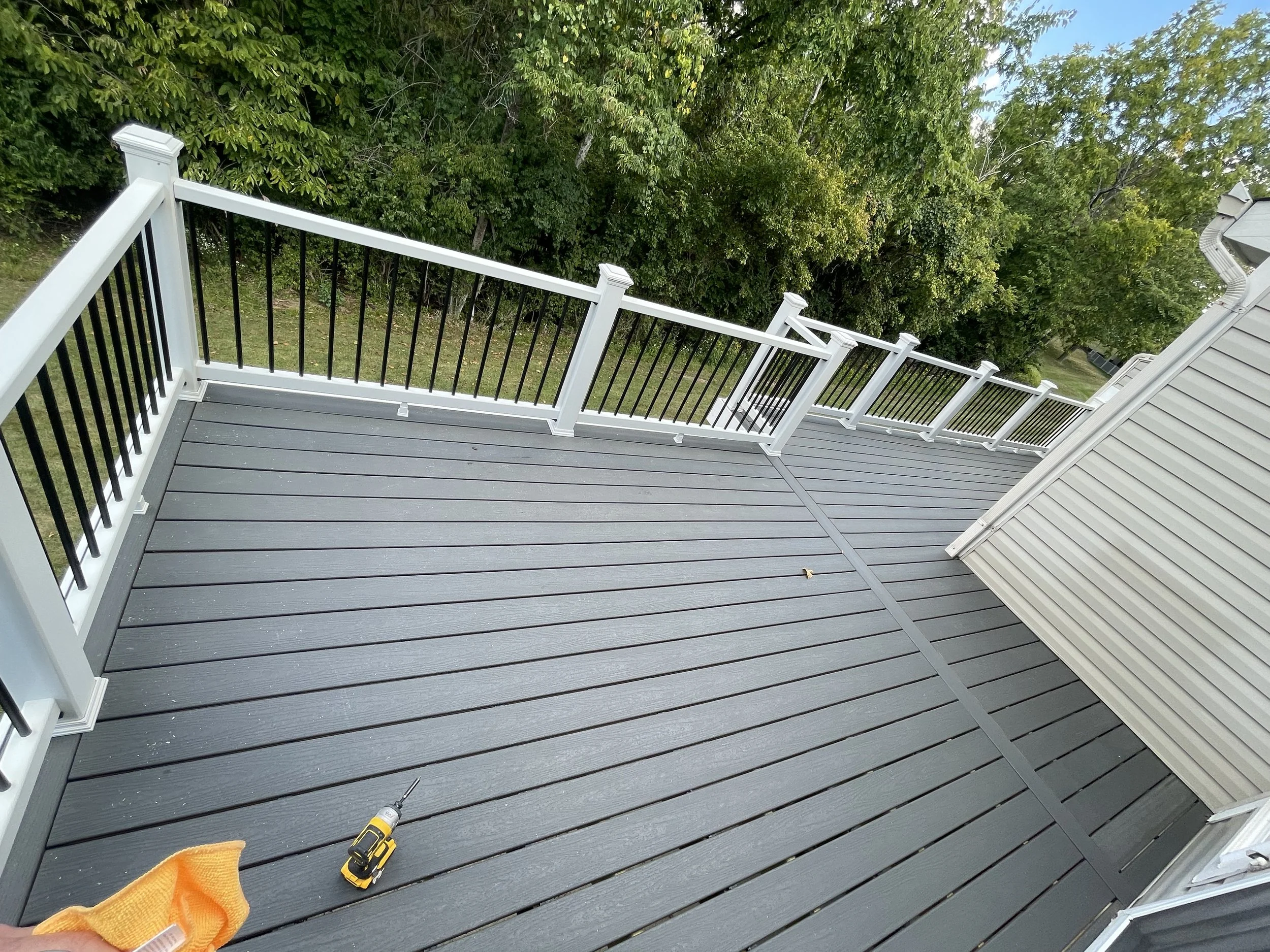 View of a newly constructed gray wooden deck with white and black railing, with a drill and a yellow cloth in the foreground, and green trees in the background.