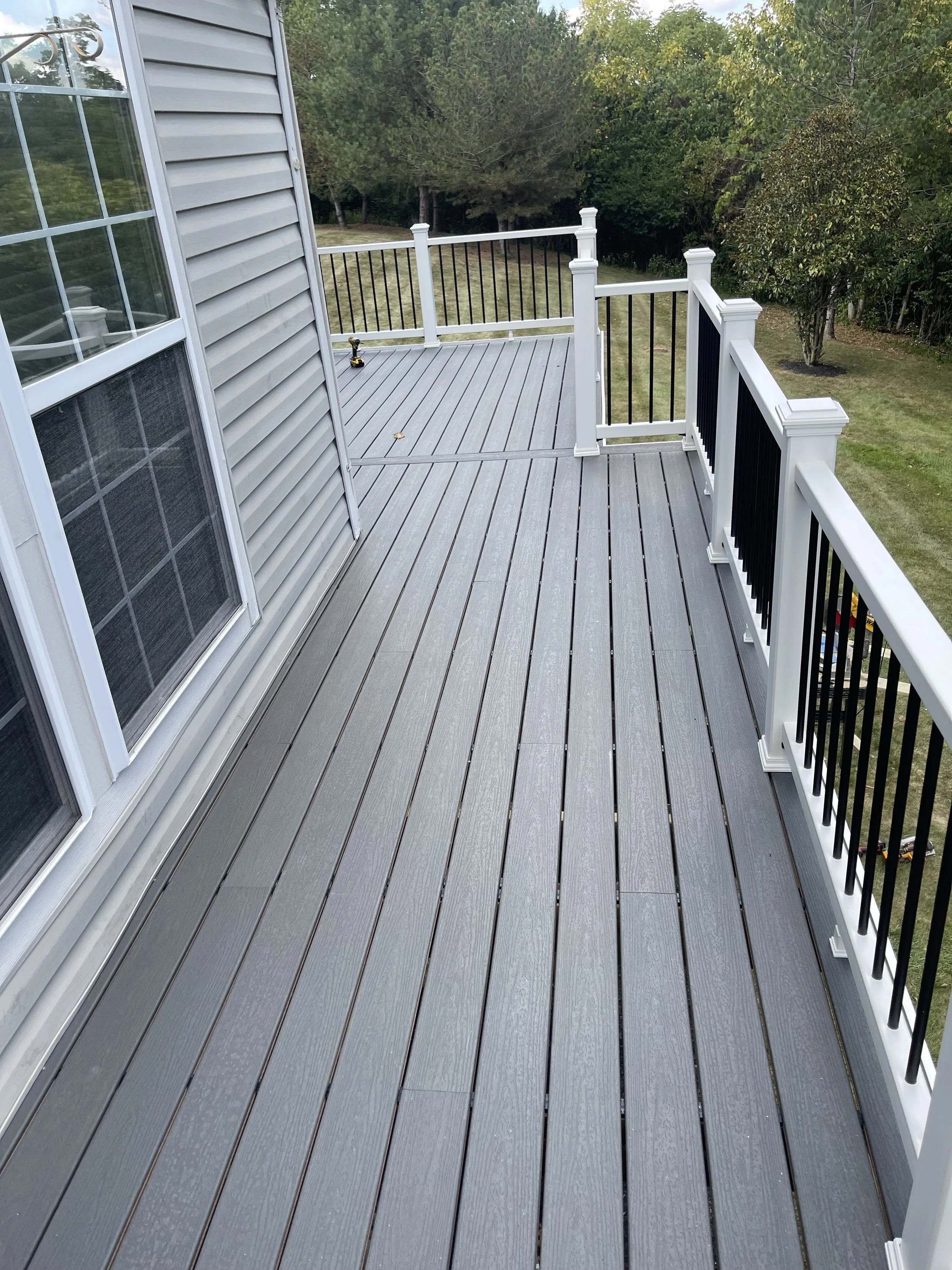 Empty gray wooden deck with white and black railing, attached to a house with light gray siding and large windows.