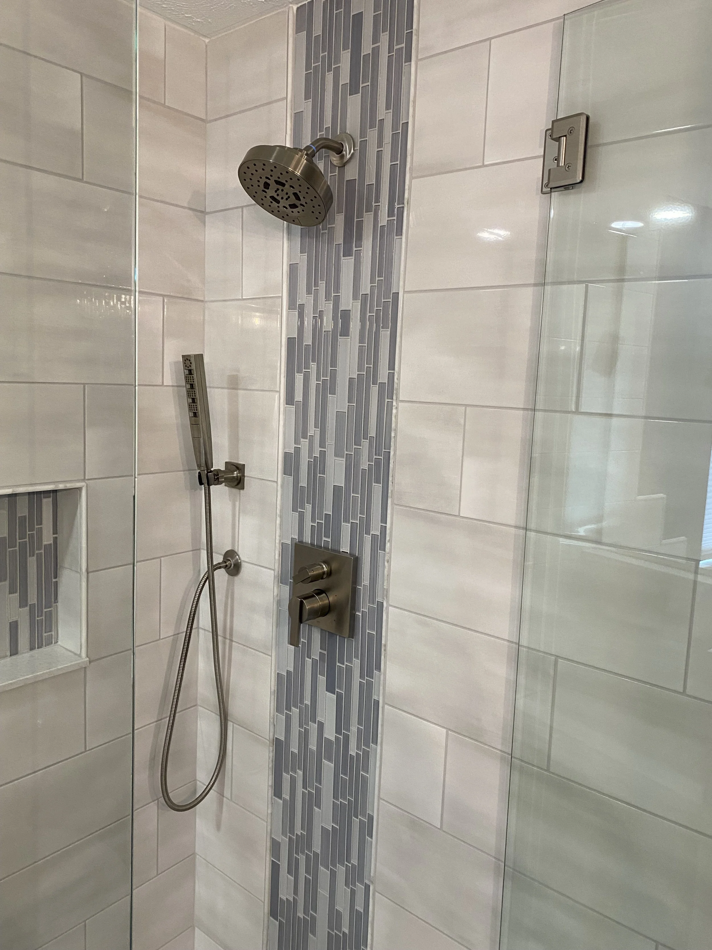 A modern shower with a rain shower head, a handheld shower head, and a wall-mounted control panel, featuring light-colored tiles with a vertical strip of gray and blue mosaic tiles.