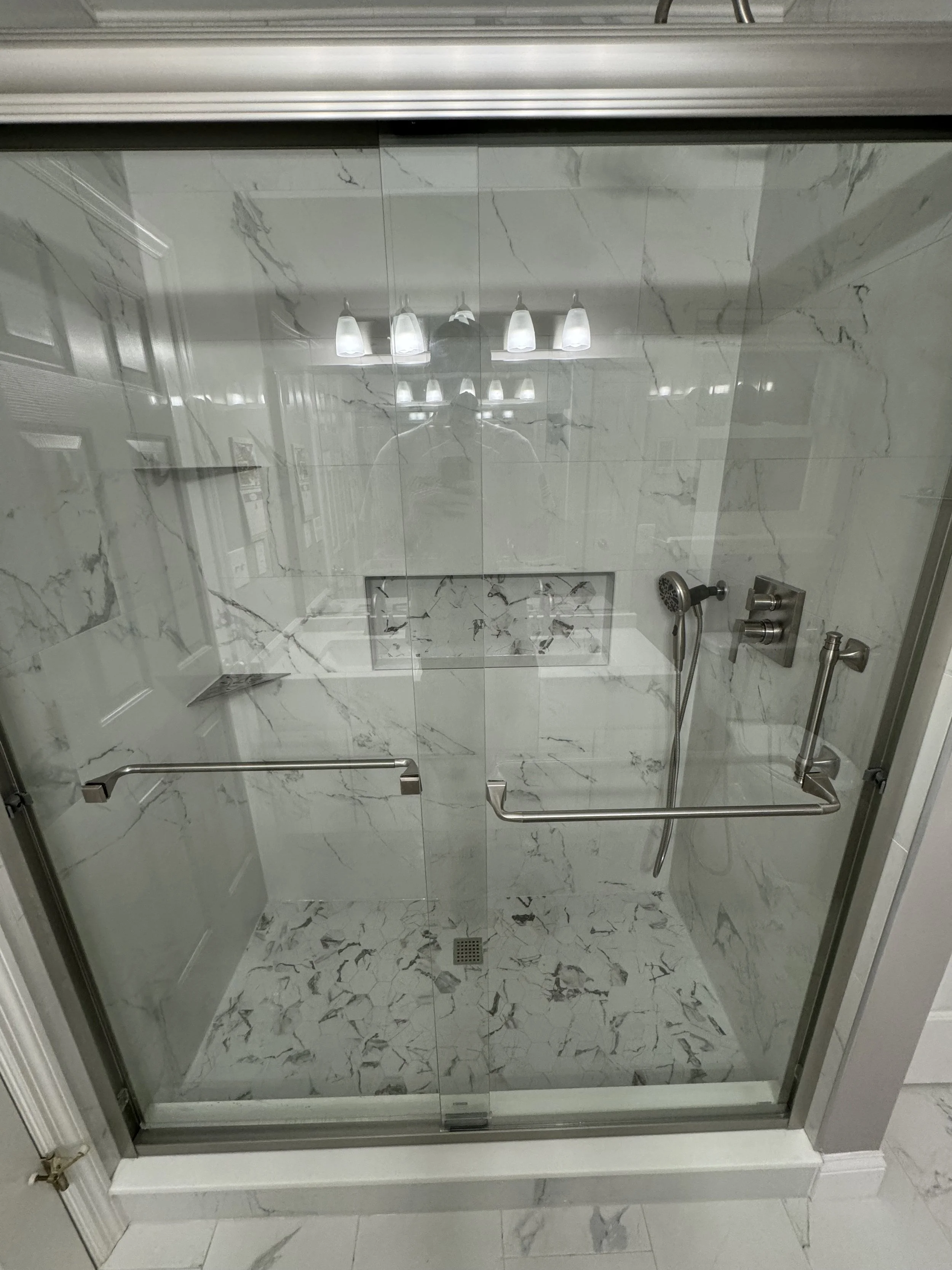 Inside a shower enclosure with glass doors, marble walls, and a built-in soap niche, featuring a handheld shower head and safety rails.