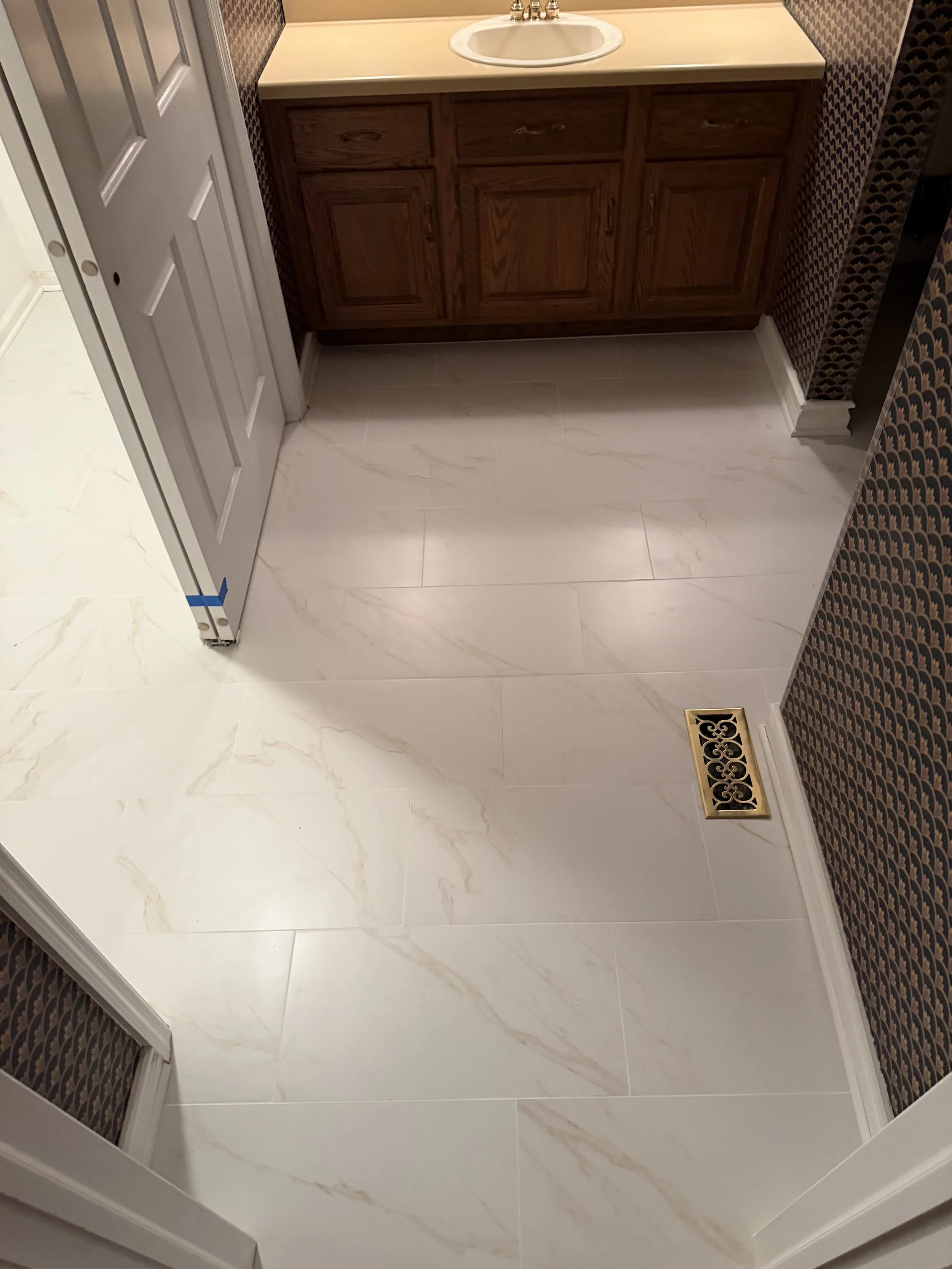 Bathroom with white marble floor, brown wooden vanity with a cream countertop and a sink, patterned wallpaper, a white door, and a gold decorative floor vent.
