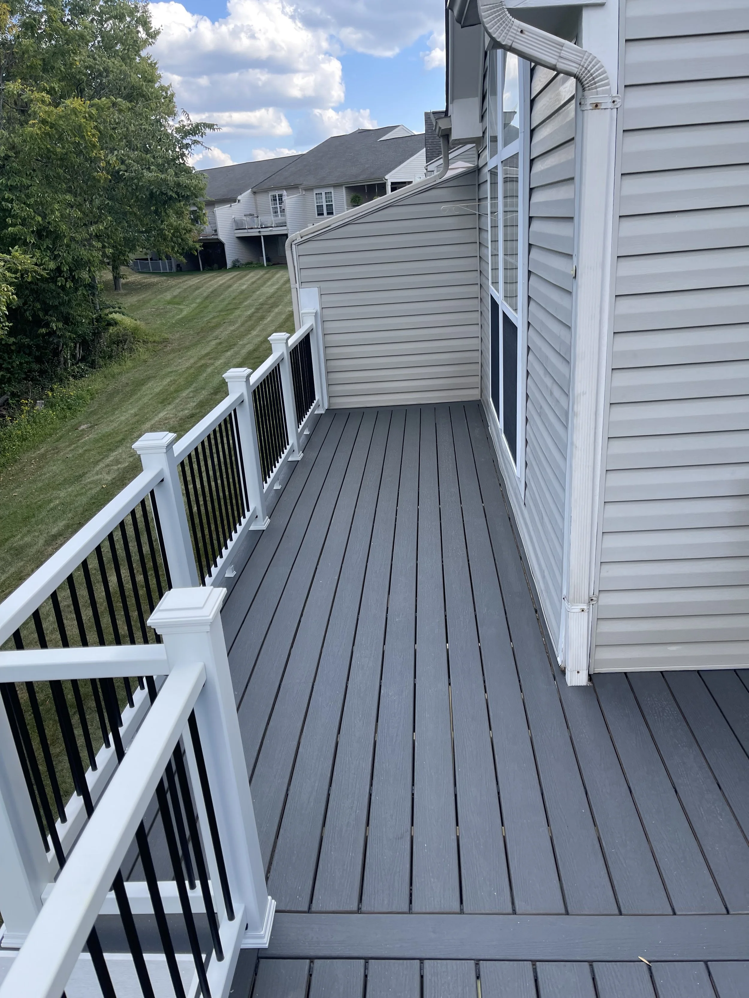 A gray wooden balcony with white and black railing, attached to a house with white siding, overlooking a grassy backyard with neighboring houses in the distance.