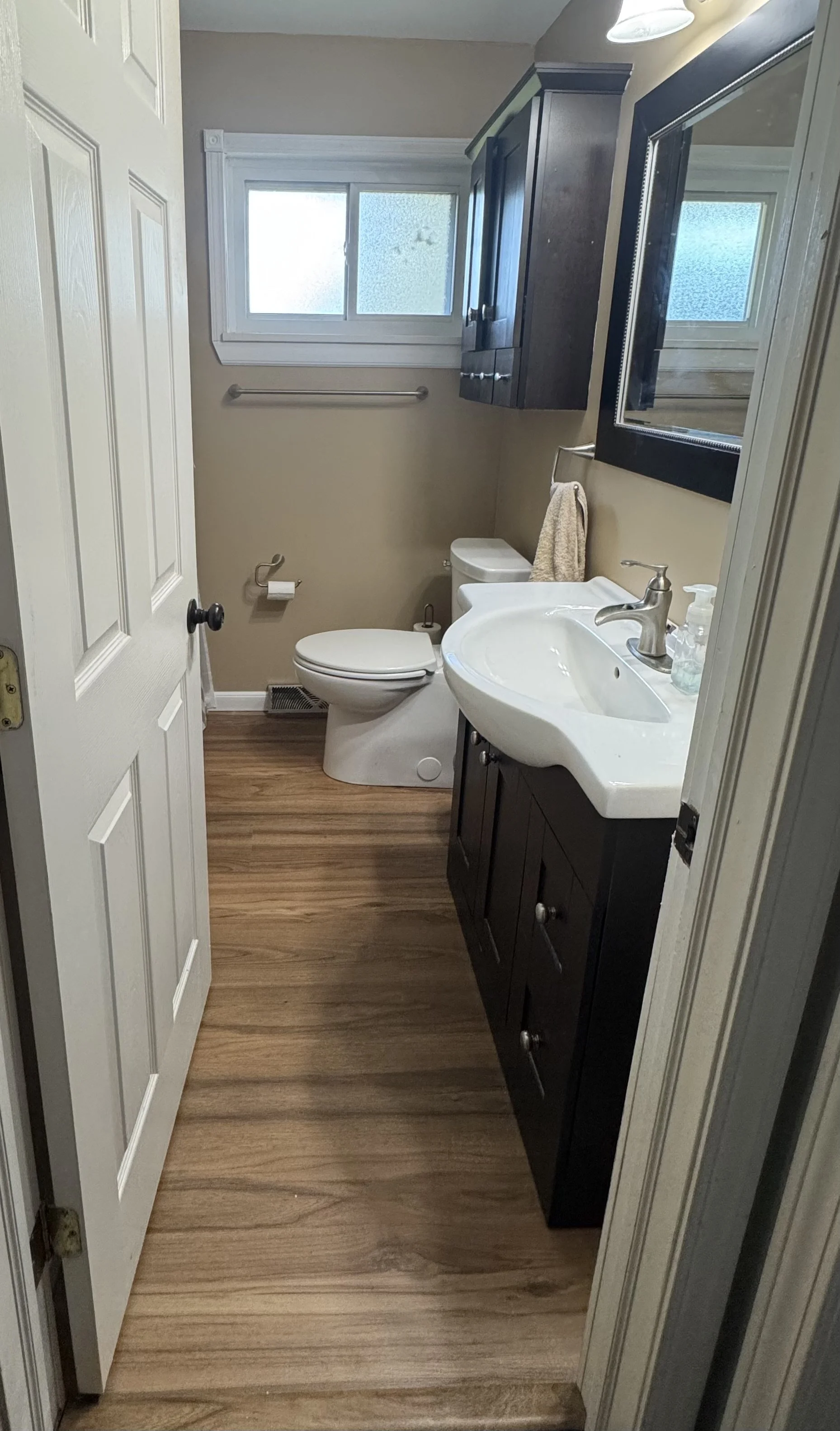 Small bathroom with wood laminate flooring, beige walls, and a white door. Features a white toilet, dark wood vanity with a curved white sink, large mirror, and dark wood upper cabinet. A frosted window is above the toilet, and a silver towel bar is 