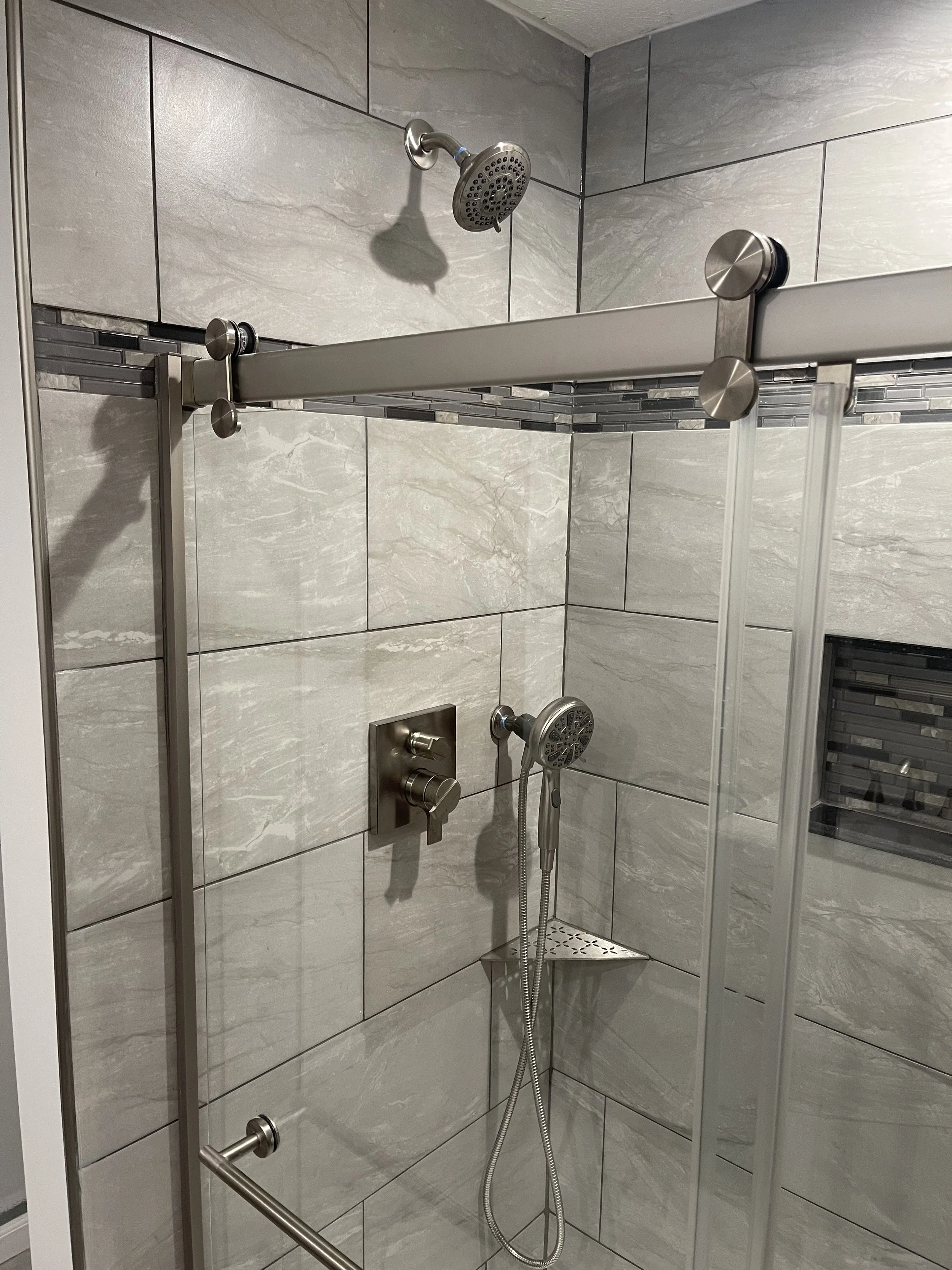 Walk-in shower with gray tiled walls, a rainfall showerhead, a handheld showerhead, and a glass sliding door.