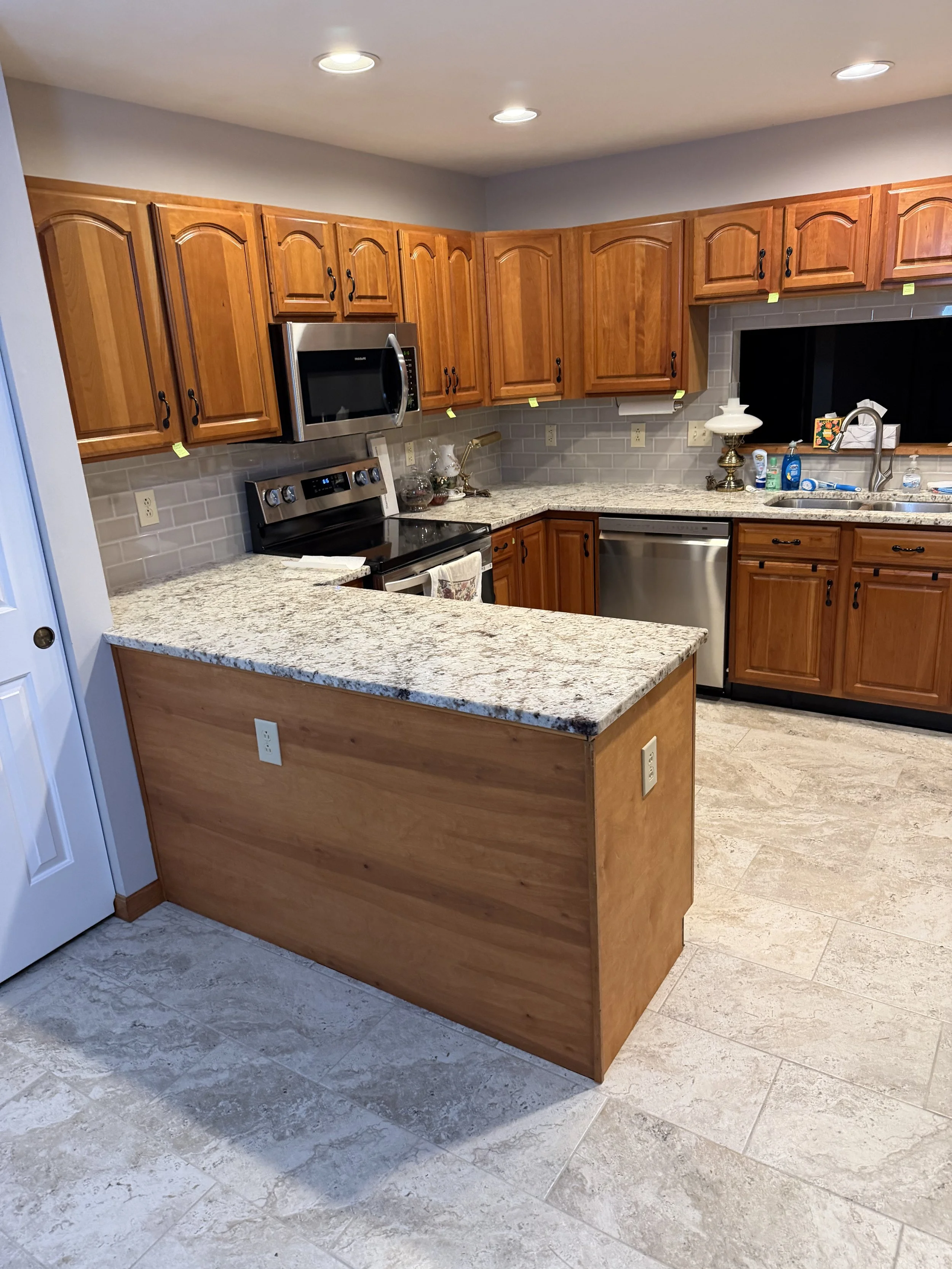 Kitchen with wooden cabinets, granite countertops, stainless steel appliances, and tiled floor. The kitchen island has electrical outlets, and the window above the sink has a black curtain.