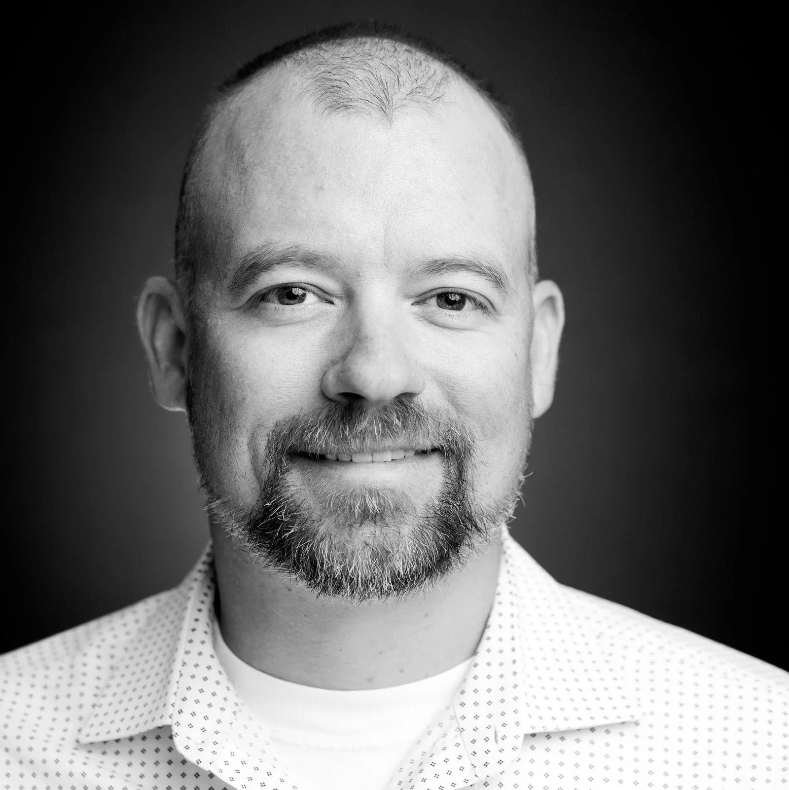 Black and white portrait of a man smiling, with a beard and short hair, wearing a collared shirt with a subtle pattern.