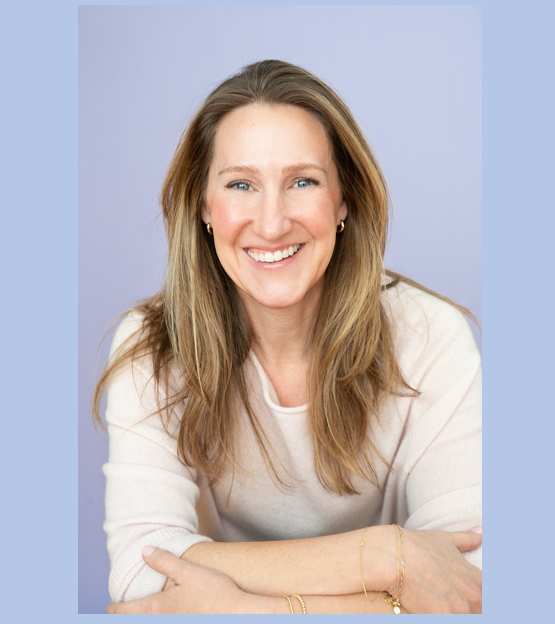 A woman with long light brown hair smiling, wearing a cream sweater and gold jewelry, against a light purple background.