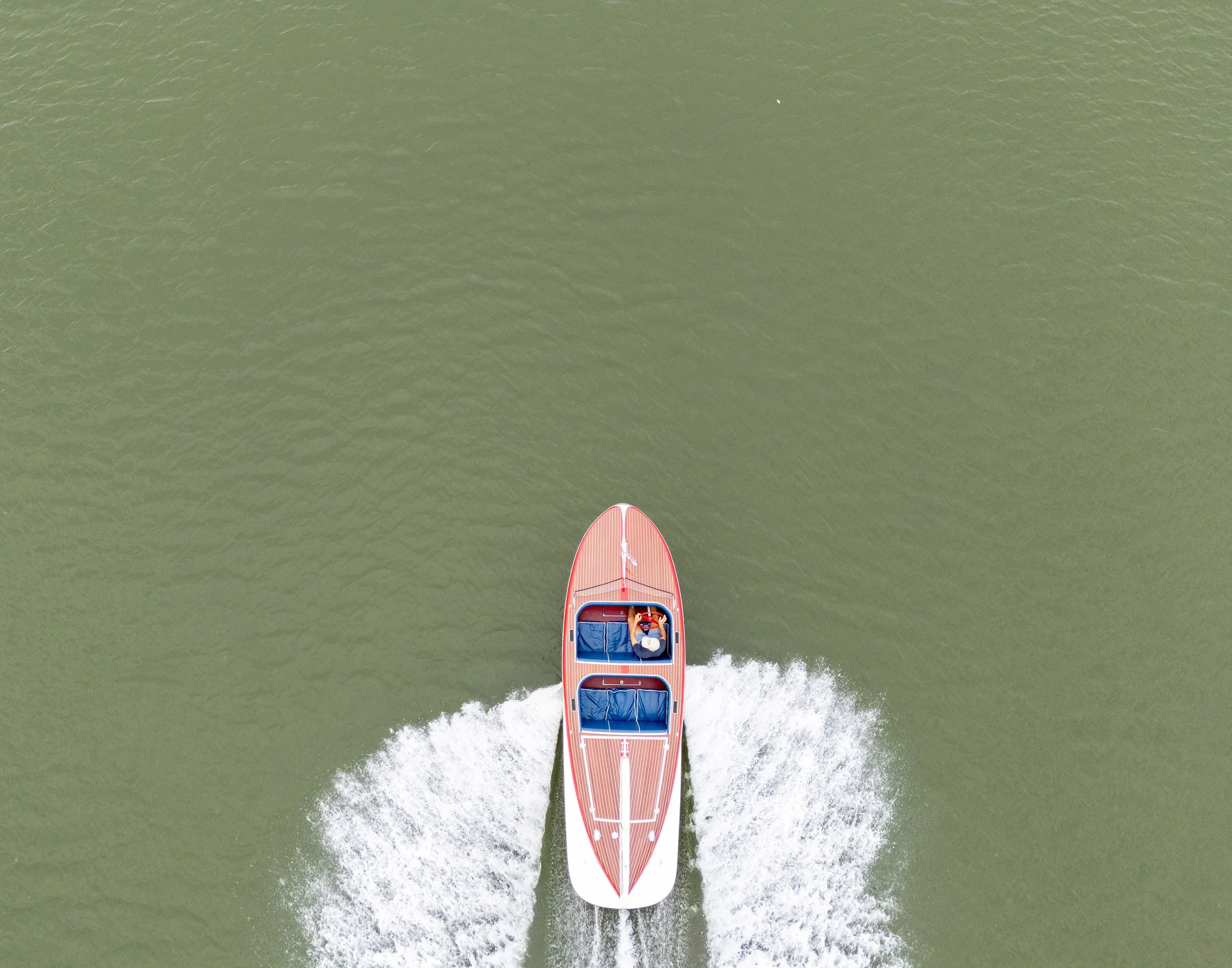 Overhead view of a small red and white motorboat speeding on green water, creating a wake behind it.