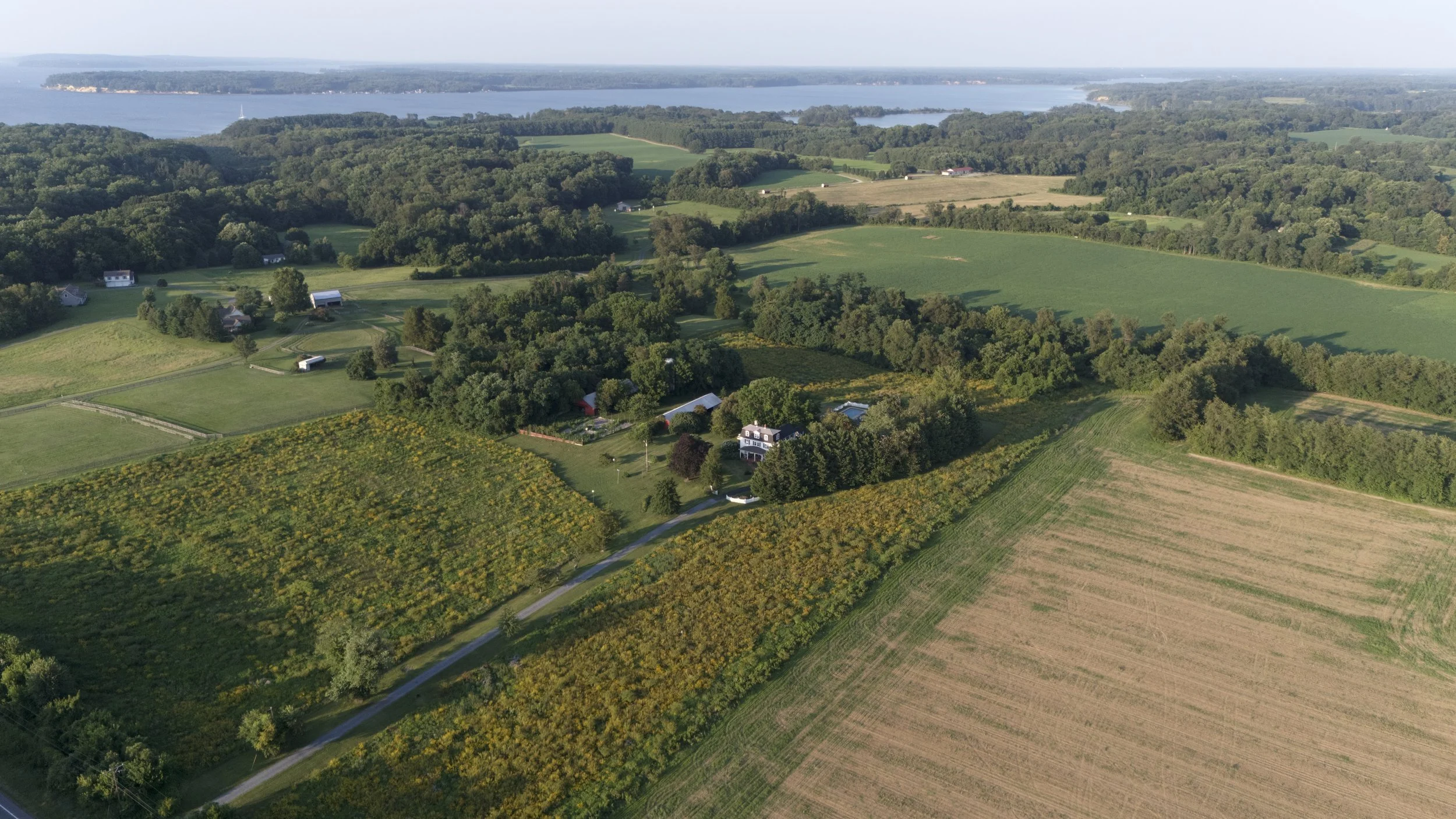 Aerial view of a rural landscape with fields, wooded areas, scattered houses, and a large body of water in the background.