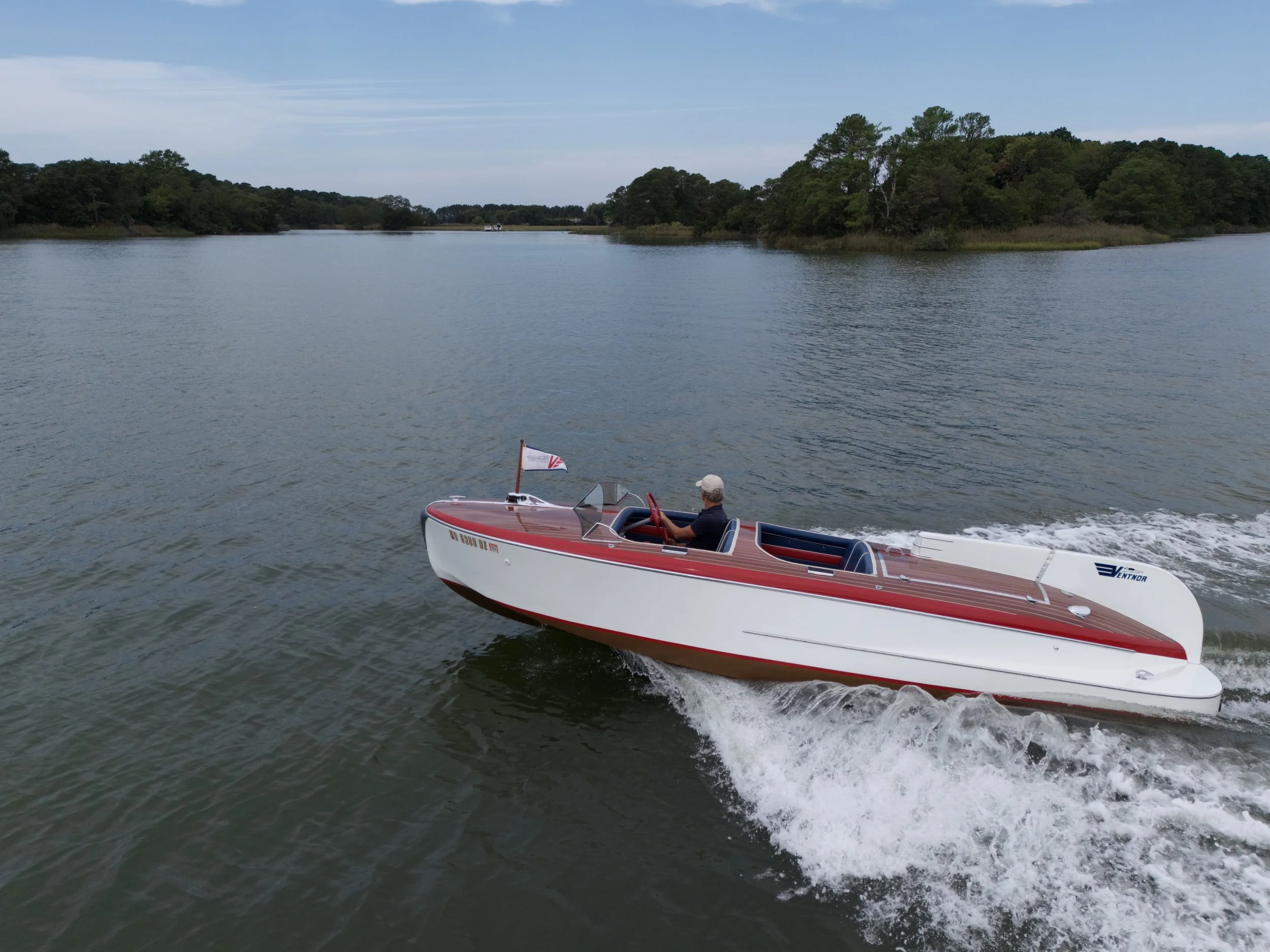 Man driving a red and white speedboat on a calm body of water, with trees and a cloudy sky in the background.