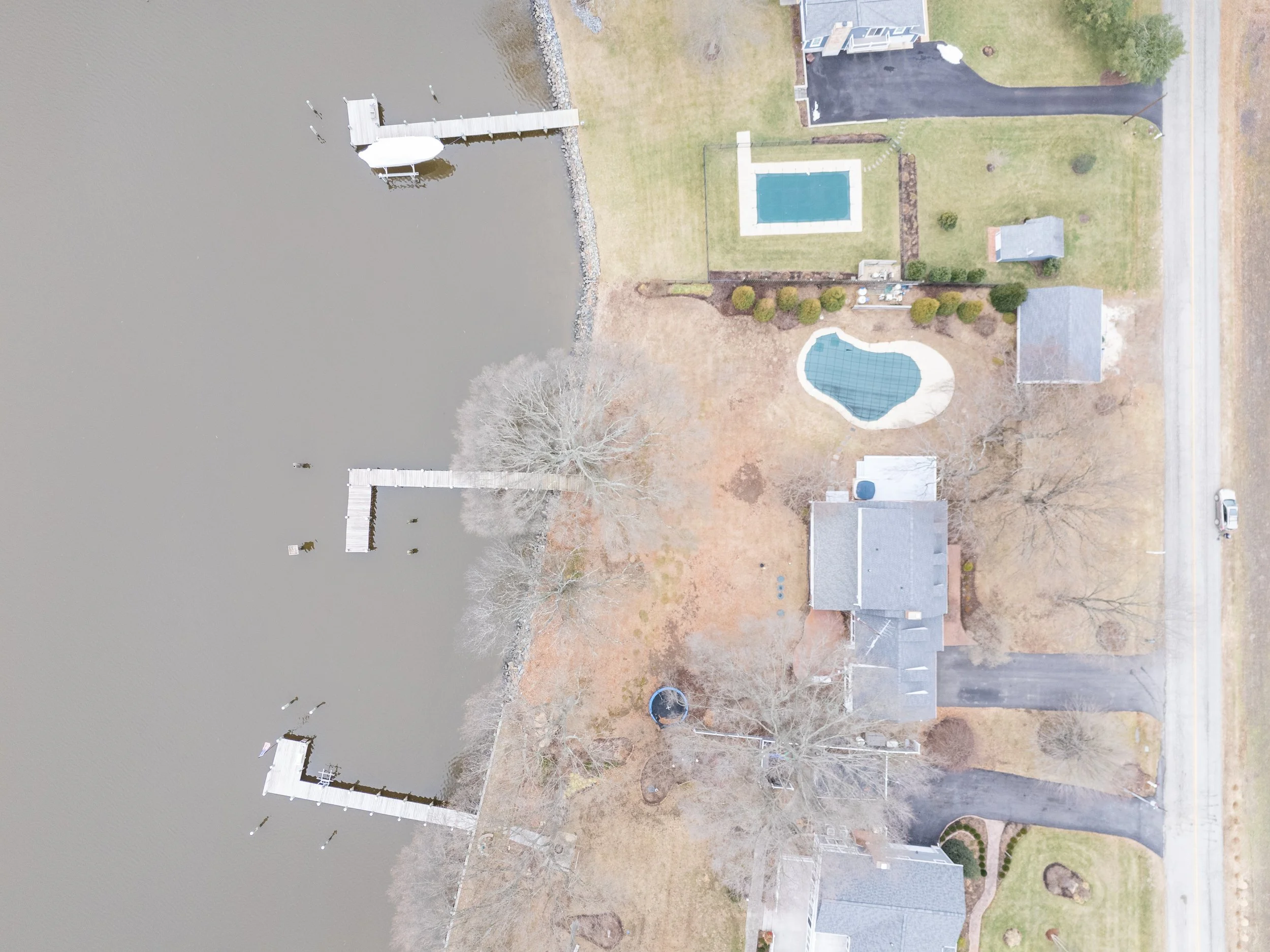 Aerial view of a residential backyard with a dock extending into a large body of water, two swimming pools, a lawn, trees, and neighboring houses.