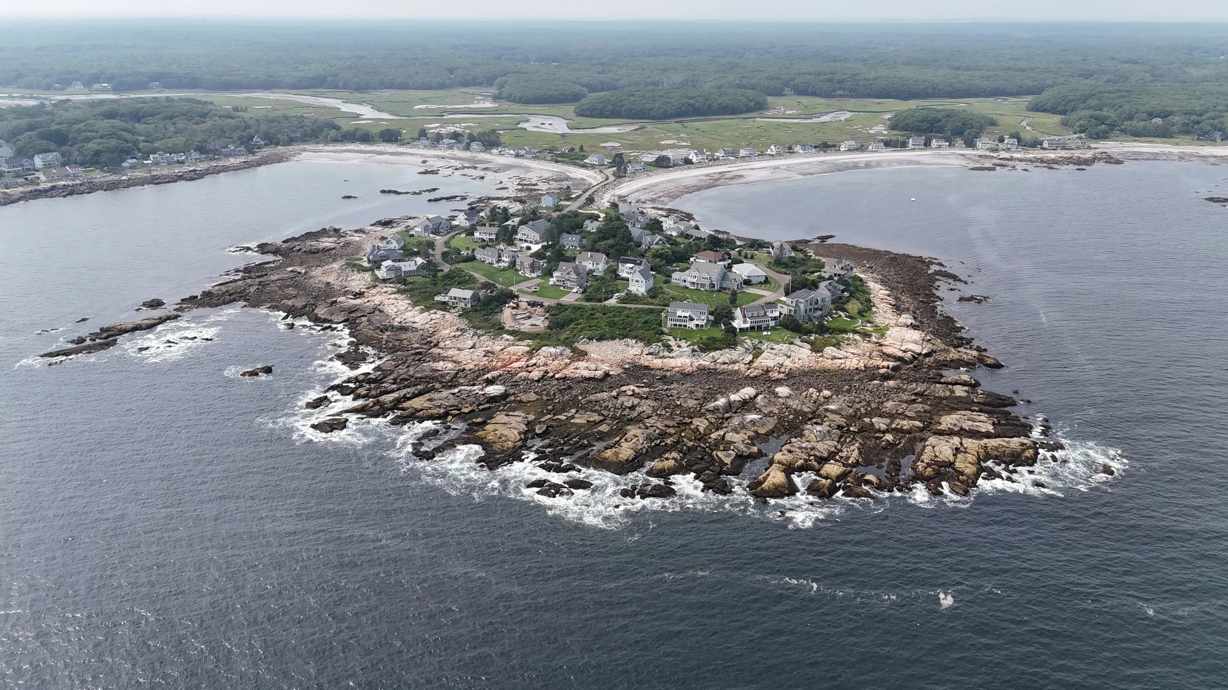 Aerial view of a coastal town with houses on a rocky peninsula surrounded by water, with a beach and green fields in the background.