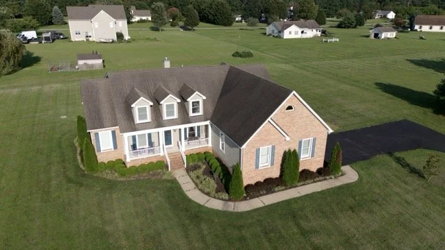 An aerial view of a single-family brick house with a white porch, surrounded by a well-kept lawn and shrubbery, with several other houses in the background.