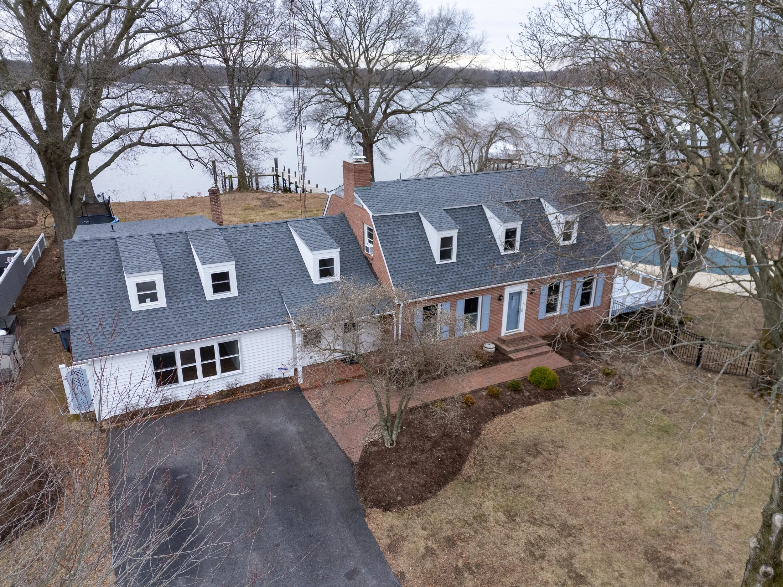 An aerial view of a house by a lake, featuring white and brick sections, dormer windows, a driveway, and leafless trees in a winter setting.