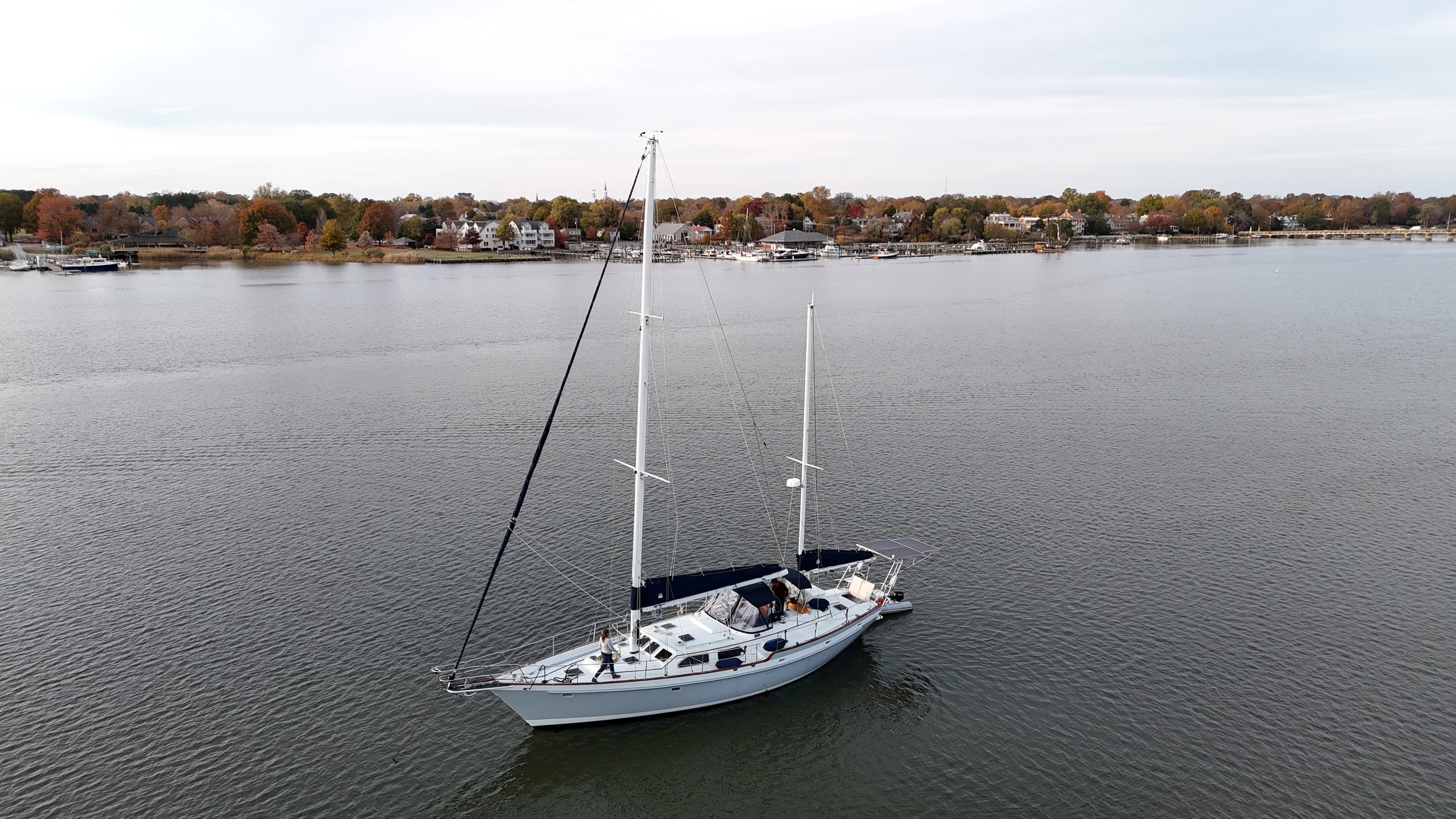 A sailboat floating on a calm river with a town and trees in the background, under a cloudy sky.