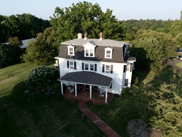 A large white, three-story house with a black roof, surrounded by trees and greenery, with a brick walkway leading to the front porch.