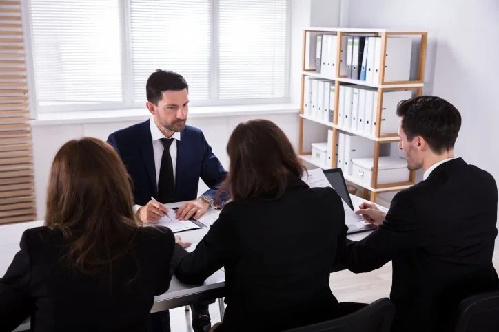 Business meeting with four people in formal attire sitting around a table in office, with shelves of files in background.