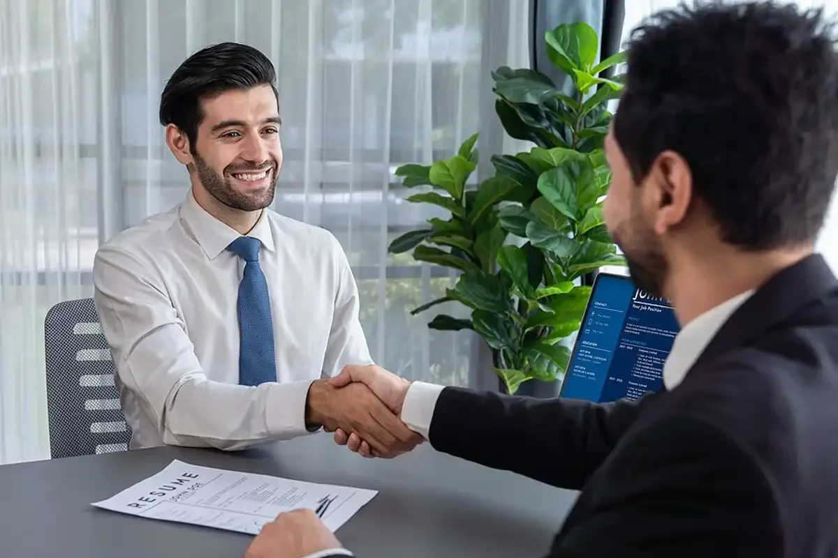 Two men in business attire shaking hands during a job interview in an office with large windows and green plants, with a resume and a resume application on the table.